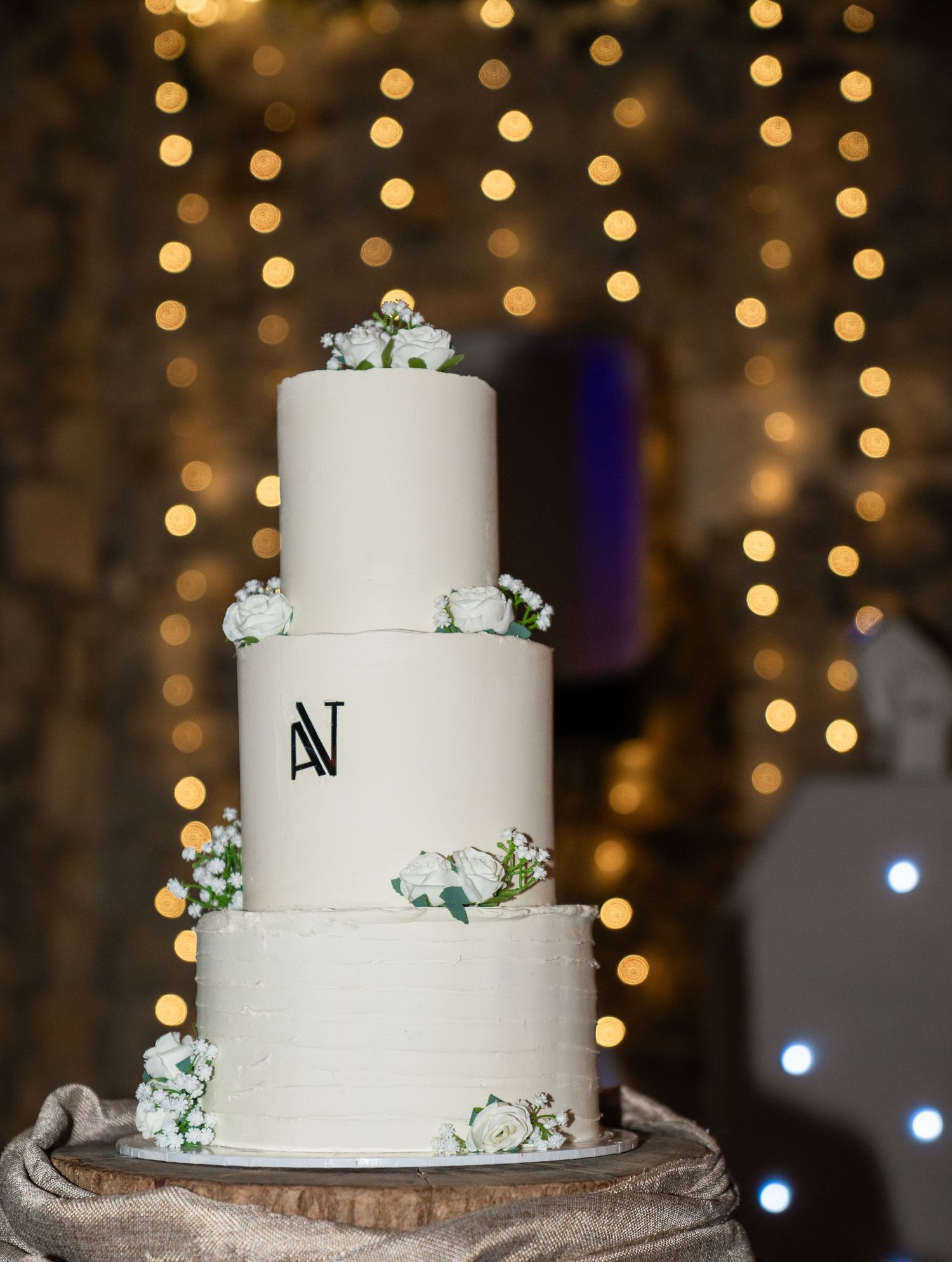 A white three-tiered wedding cake with eucalyptus accents sits on a rustic wooden stand against a bokeh light background.