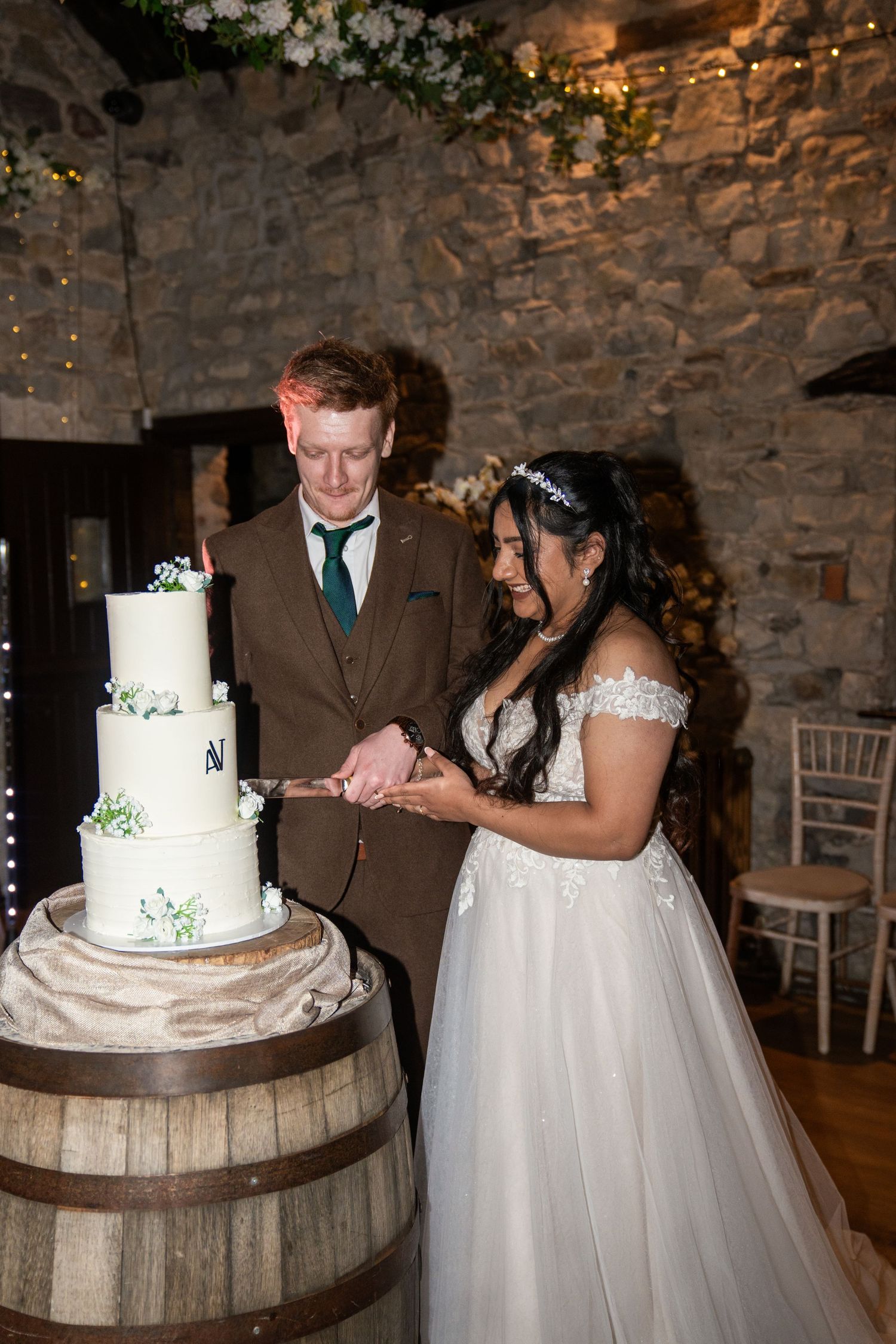 A wedding couple cuts their elegant three-tiered white cake displayed on a rustic wooden barrel in a stone-walled venue.