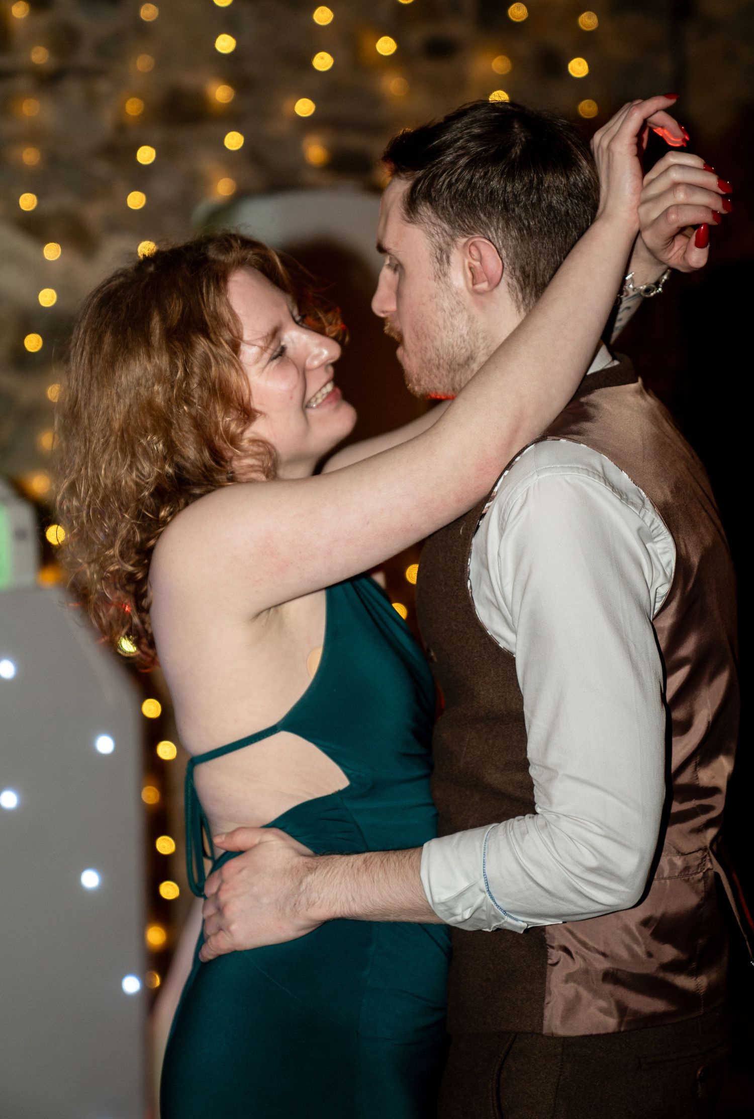 A couple shares a romantic dance together under warm twinkle lights at an evening celebration.