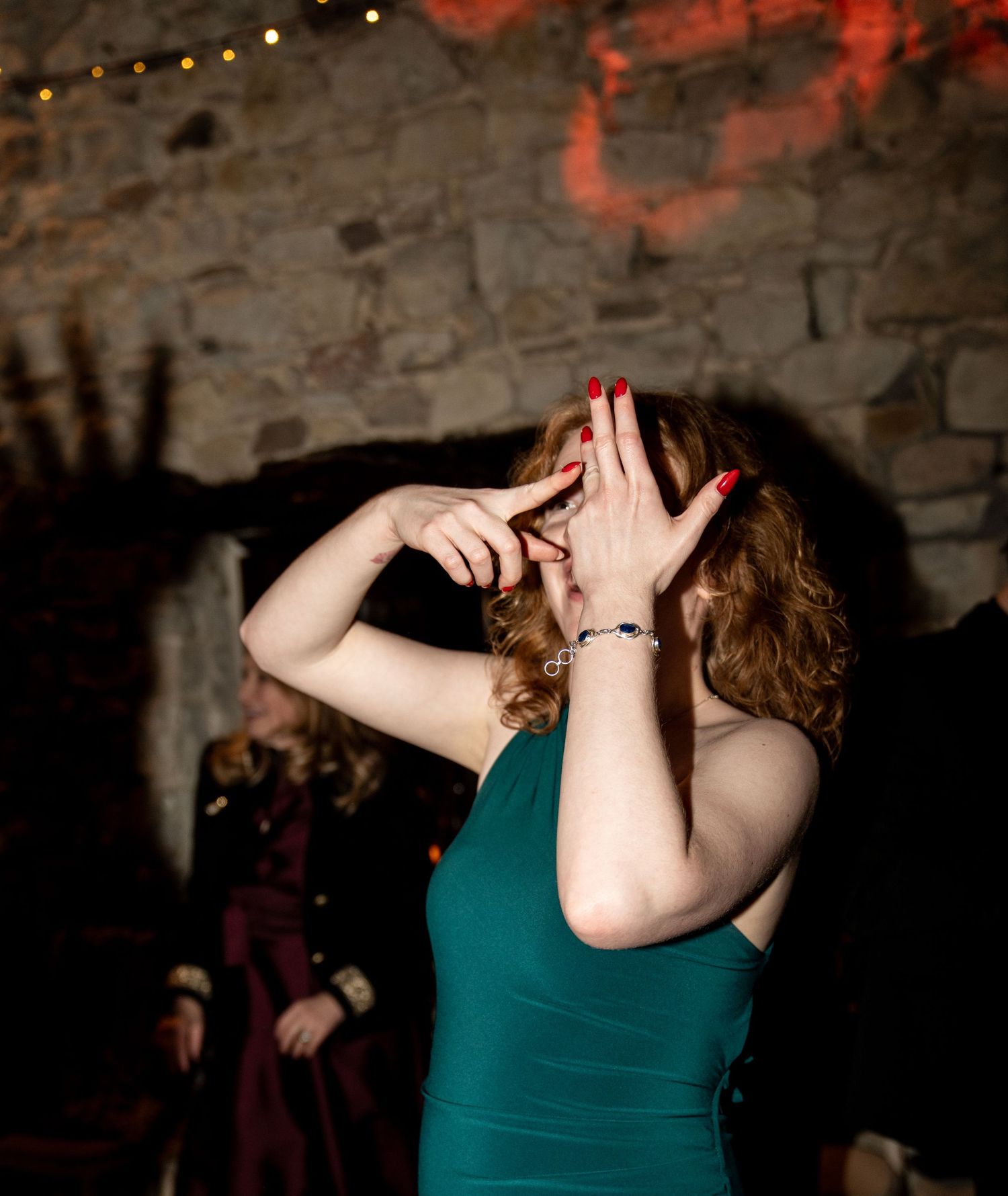 A person in a teal tank top dances expressively in a dimly lit space with brick walls and string lights.