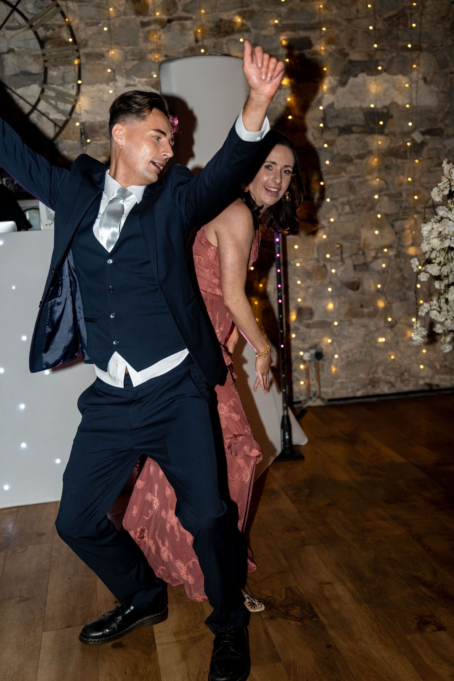 Guests dancing energetically on a wooden dance floor at a party with twinkle lights and exposed brick walls in background.