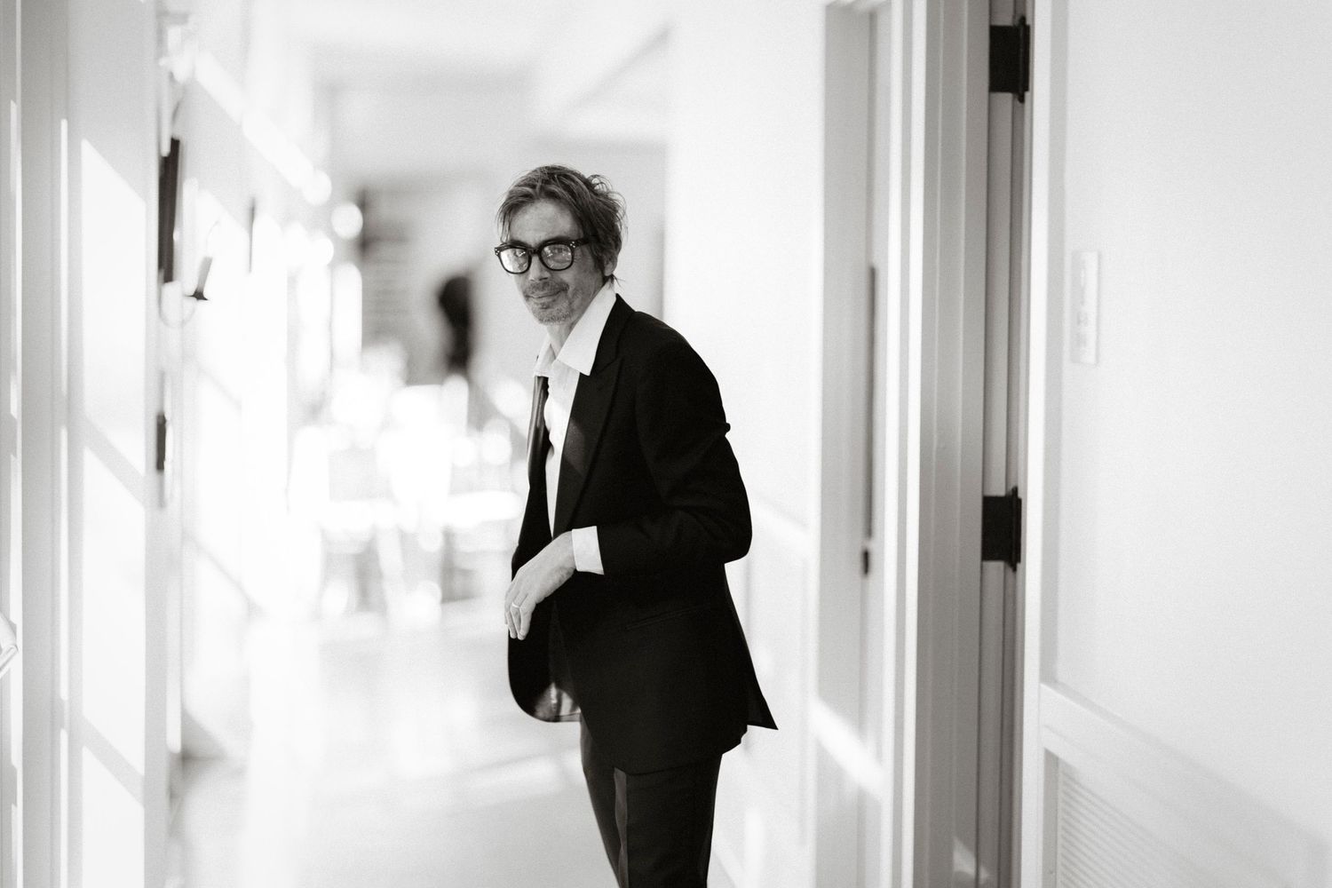 A person in a black suit and glasses walks through a bright white hallway in a black and white photograph.