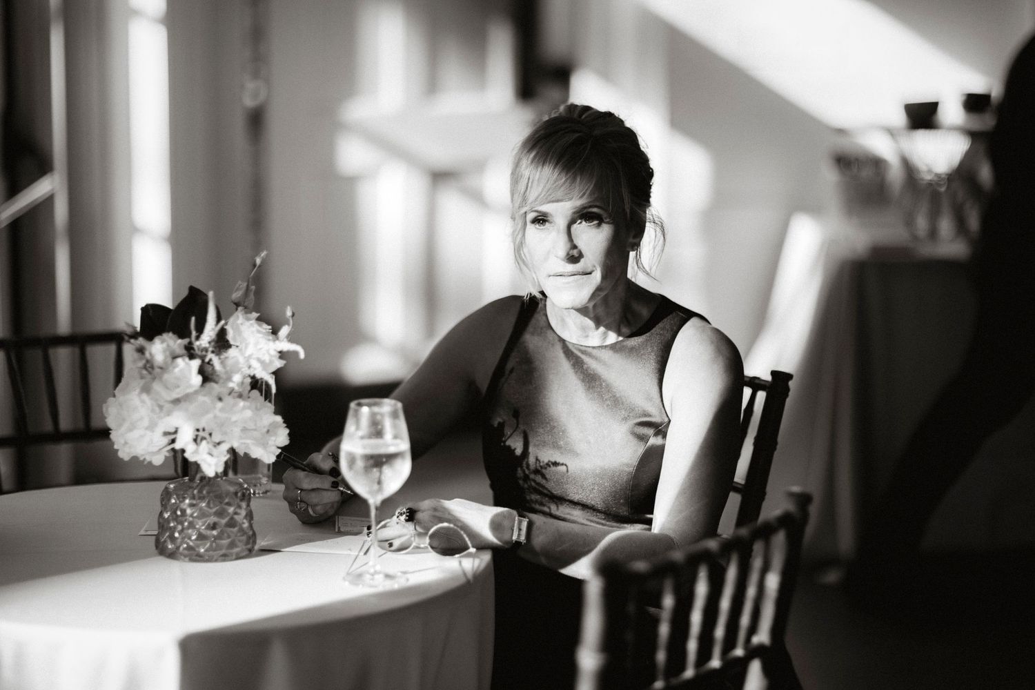 Black and white photo of someone seated at a restaurant table with wine glass and floral centerpiece in elegant setting.