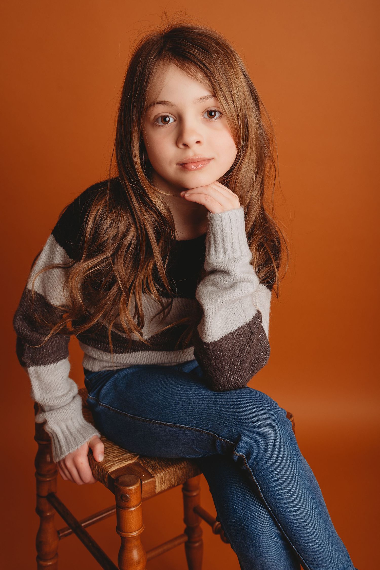Portrait in striped sweater sitting on wooden stool against orange backdrop.