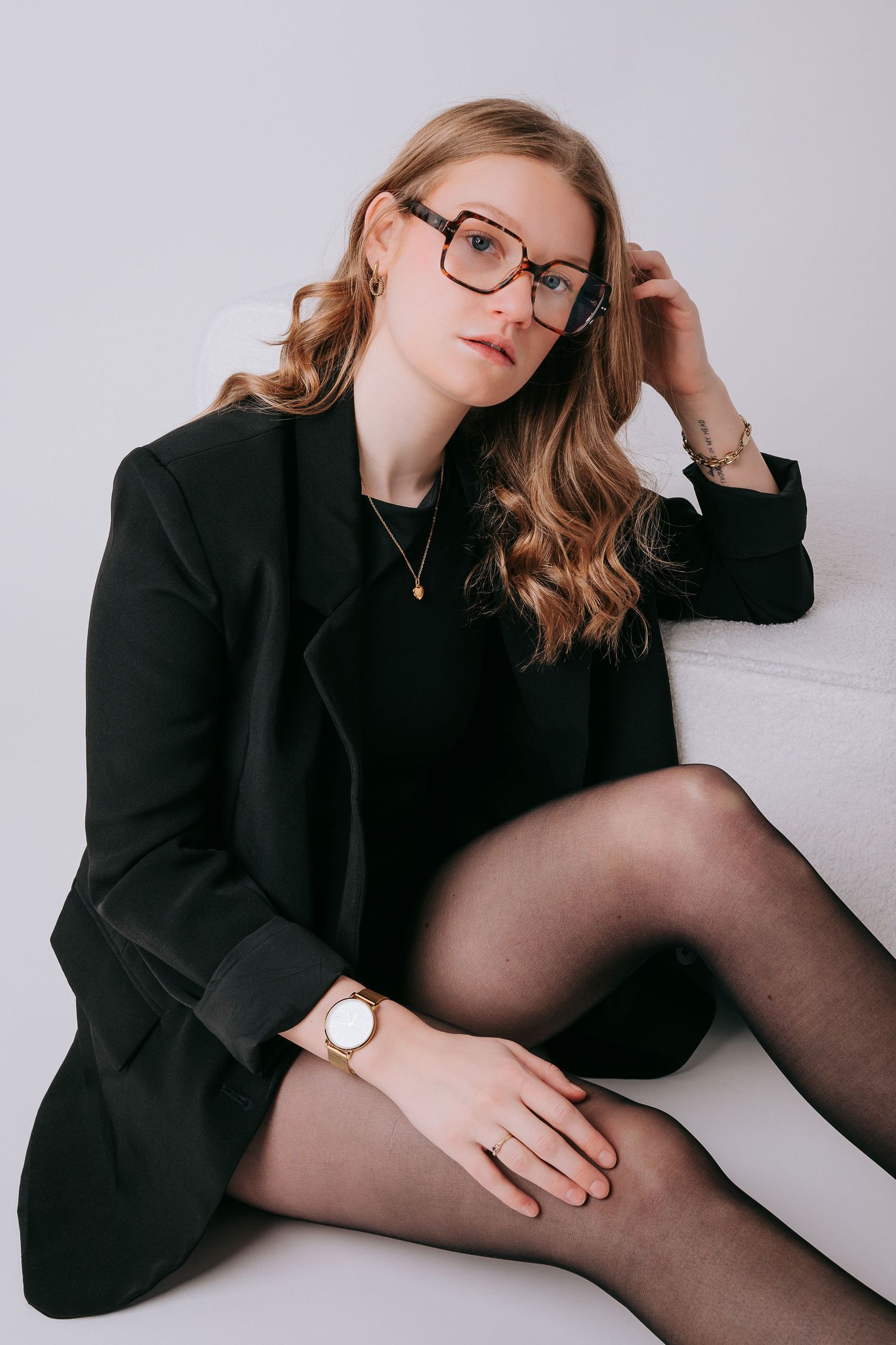 Young woman in black blazer and tights, wearing oversized glasses and gold watch, sitting elegantly against white background.