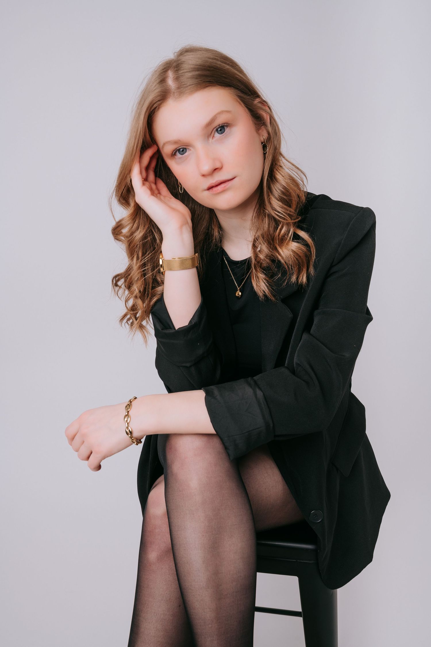 Young woman in black blazer and tights seated on stool, wearing gold bracelet and watch, with wavy blonde hair.