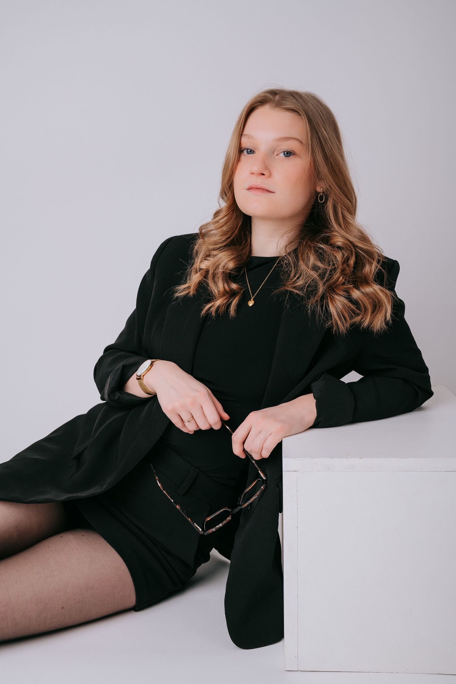 Young woman in black blazer and dress sits beside white cube, posing elegantly against a light gray background.