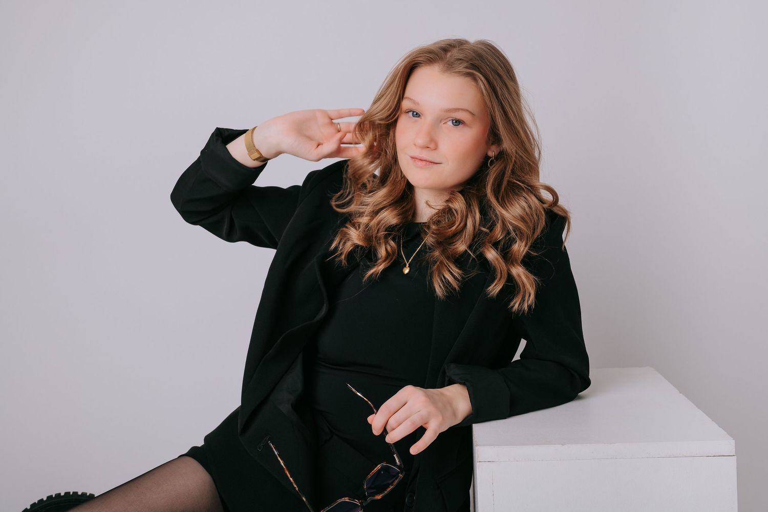 Young woman in black blazer poses confidently on white cube, holding glasses, with curly hair and gold necklace.