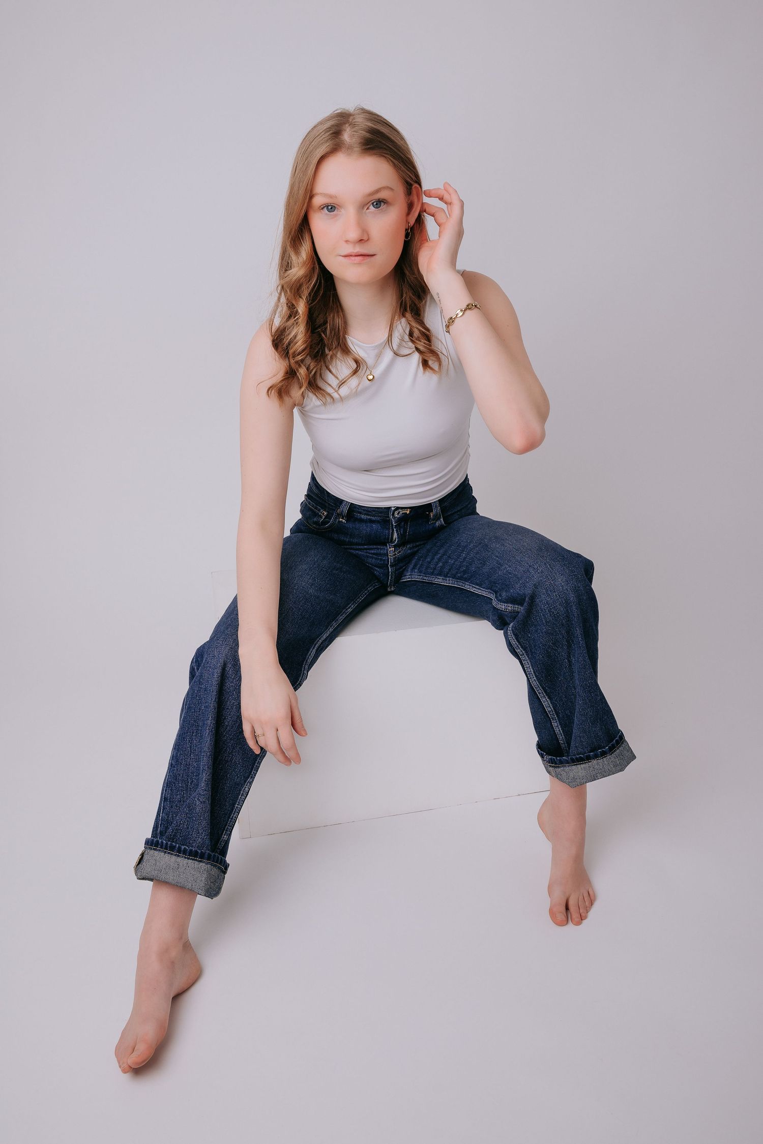 Young woman in white tank top and dark blue cuffed jeans sitting barefoot on white cube in studio.