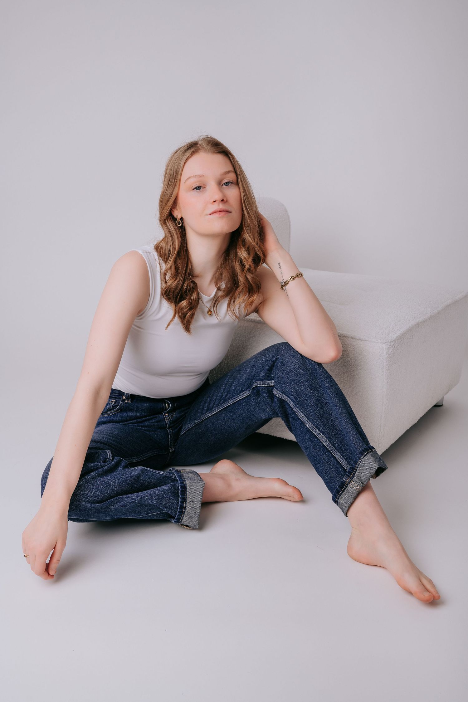 Young woman in white tank top and rolled blue jeans sitting barefoot on floor leaning against white chair.