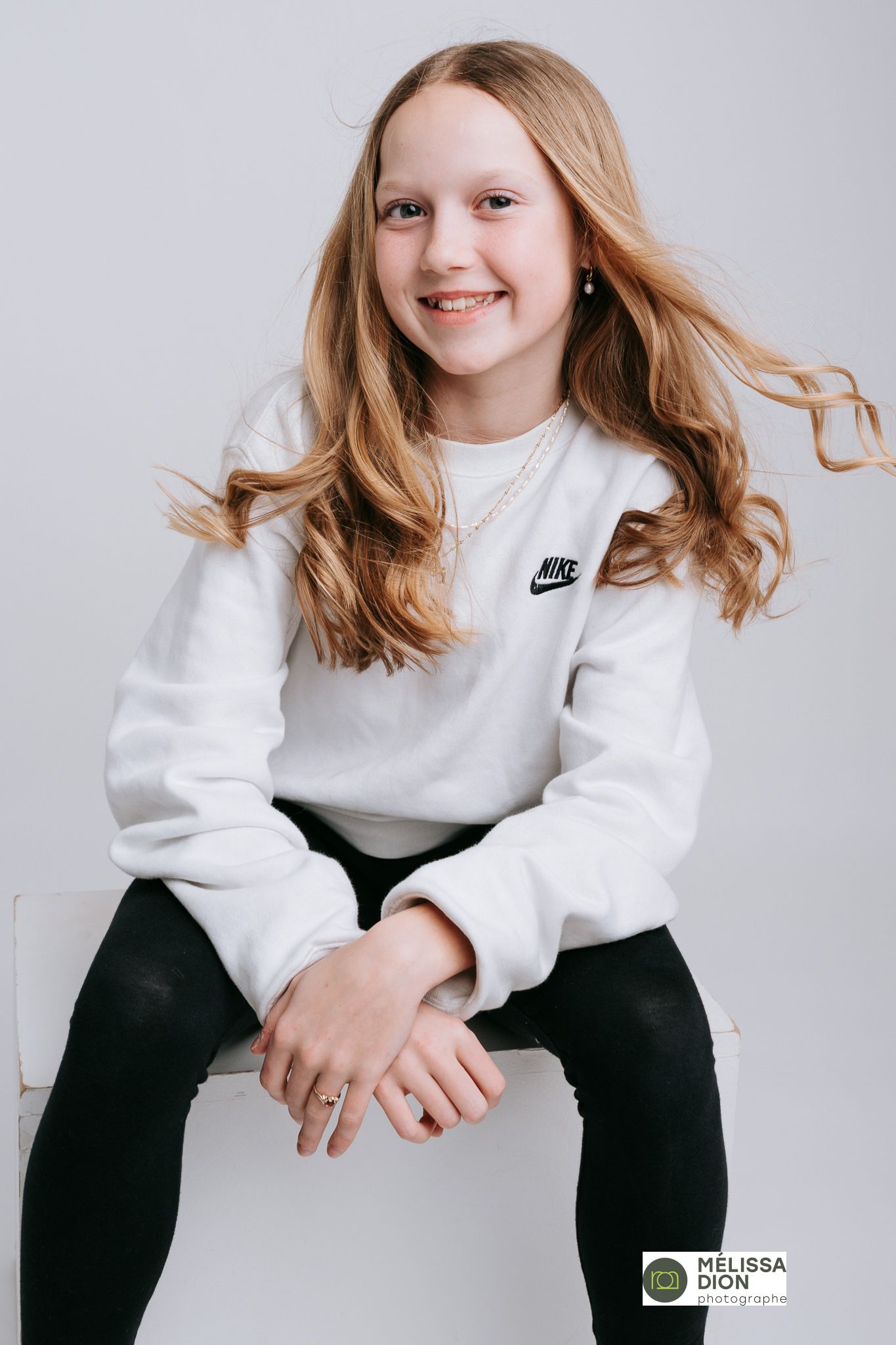 Smiling young girl with long blonde hair wearing a white Nike sweatshirt and black leggings, seated against a white background.