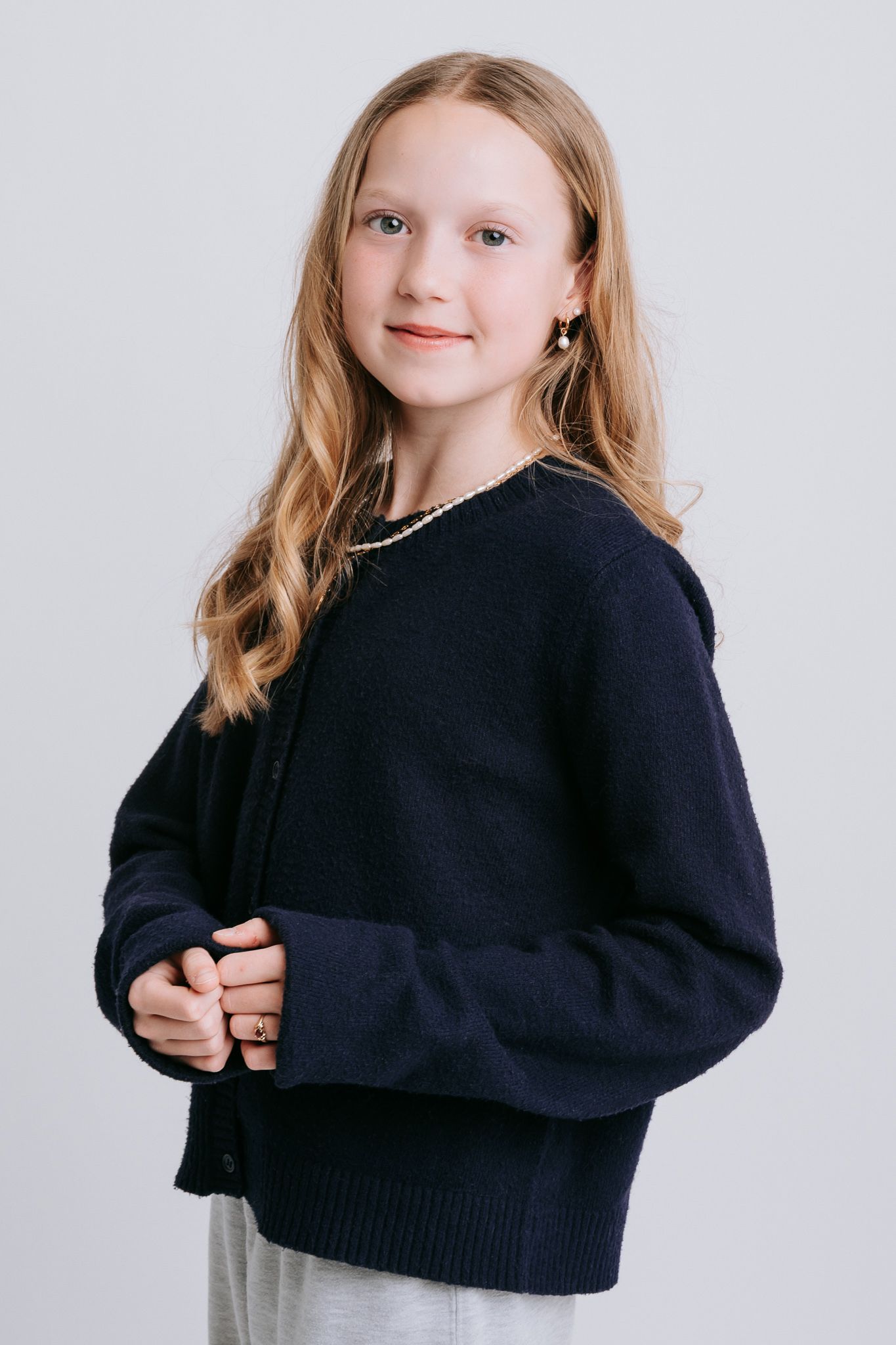 Young girl with long blonde hair wearing a navy blue knit cardigan, posing against a light gray background.