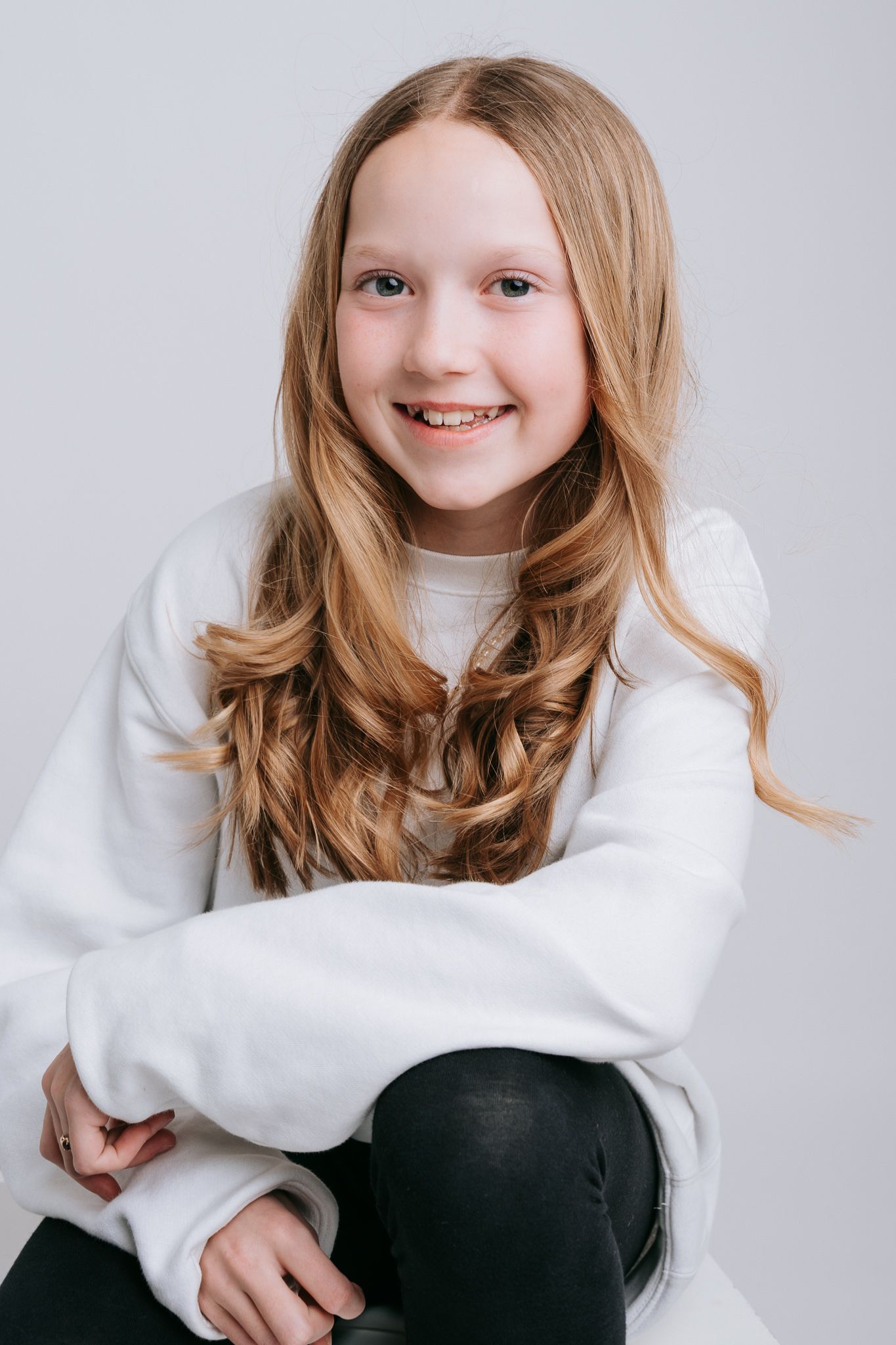 Smiling young girl with long wavy blonde hair wearing a white sweatshirt and black leggings on gray background.