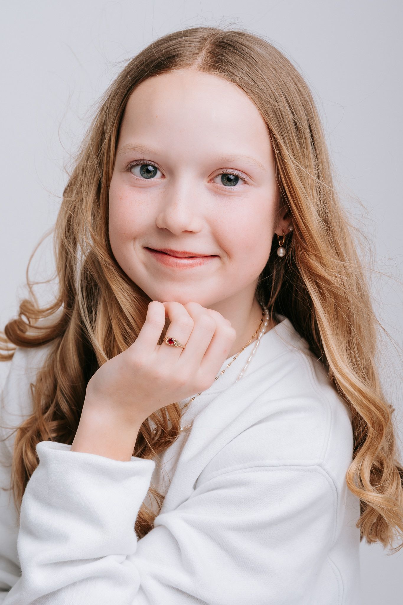 Young girl with long blonde hair and blue eyes wearing a white sweater, posing gracefully.