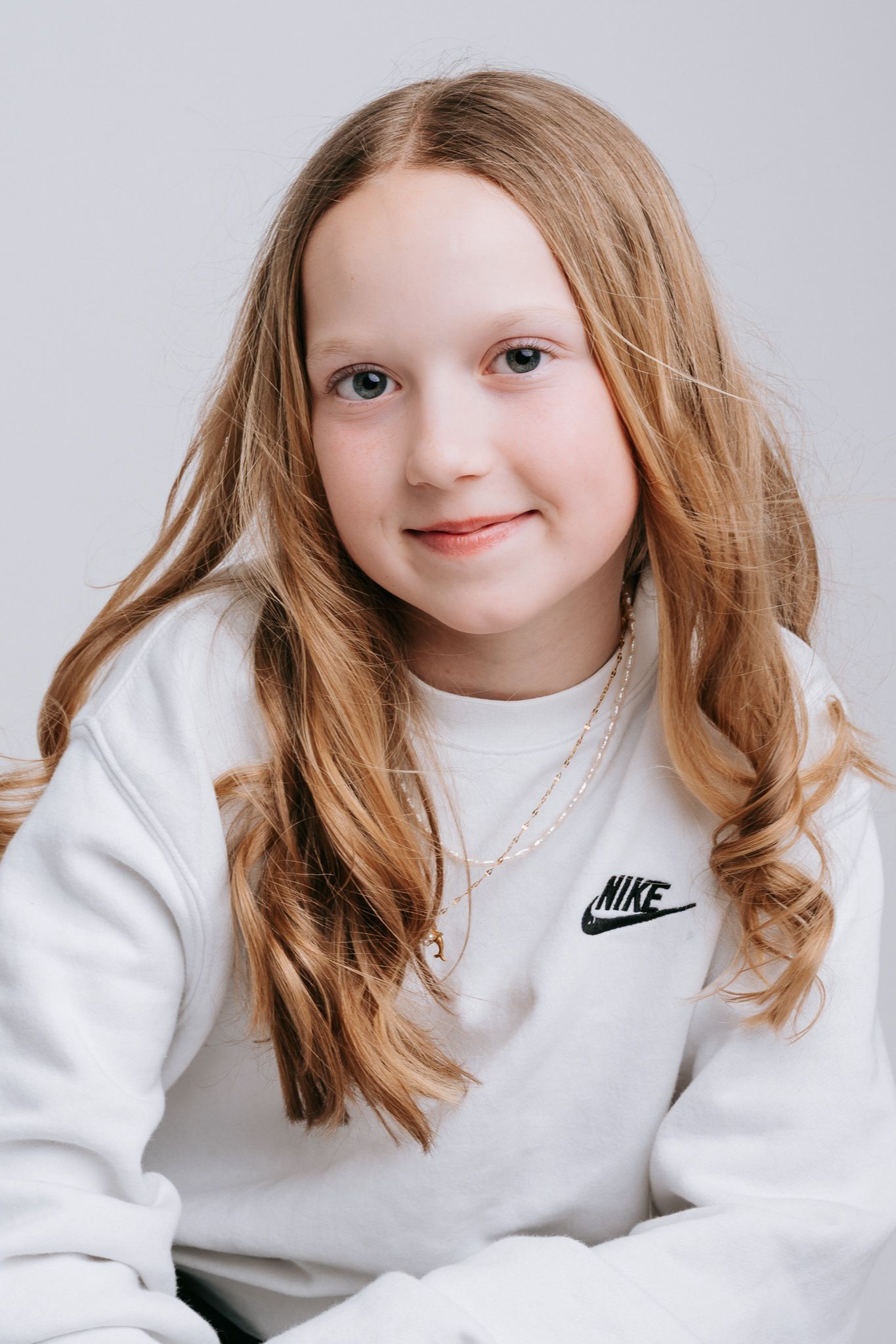 Young girl with long blonde hair wearing a white Nike sweatshirt and delicate necklace, smiling softly against a light background.