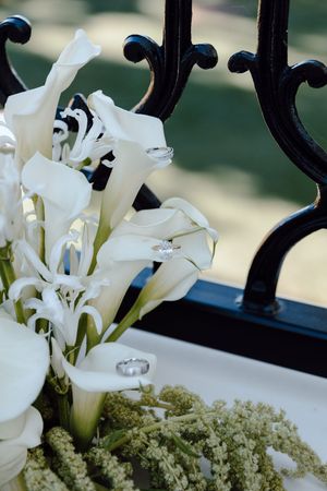 White calla lilies arranged with delicate green foliage against an ornate black wrought iron background.