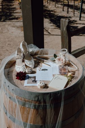 Wedding accessories including white heels, invitation, rings and flowers arranged on rustic wooden wine barrel.