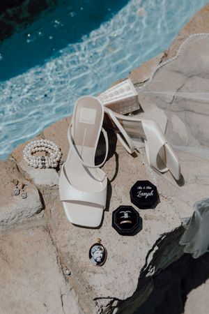 White wedding shoes and rings arranged artistically on natural stone near turquoise water in a coastal setting.