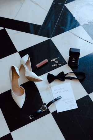 Wedding accessories including white heels, rings, and invitations arranged on a black and white checkered floor.