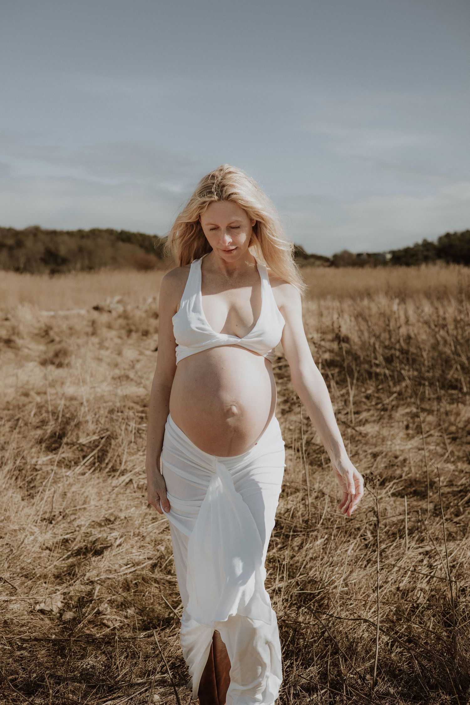 gravid kvinna sitter på en stock och kollar bortåt. på sig har hon ett vit set och bruna stövlar.