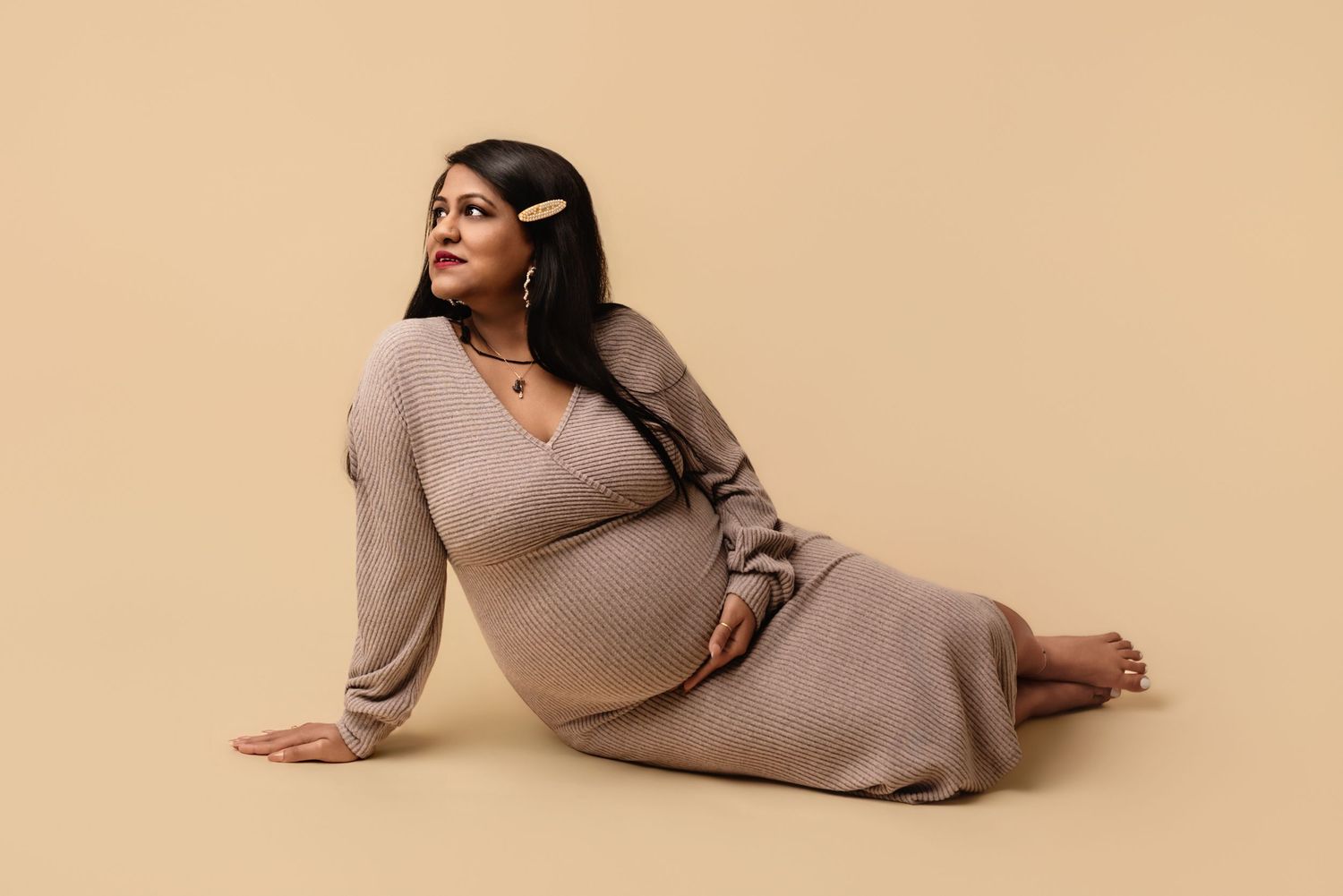 Expectant mom in fitted knit neutral dress stretches across a beige backdrop, captured by Charlottesville maternity photographer.