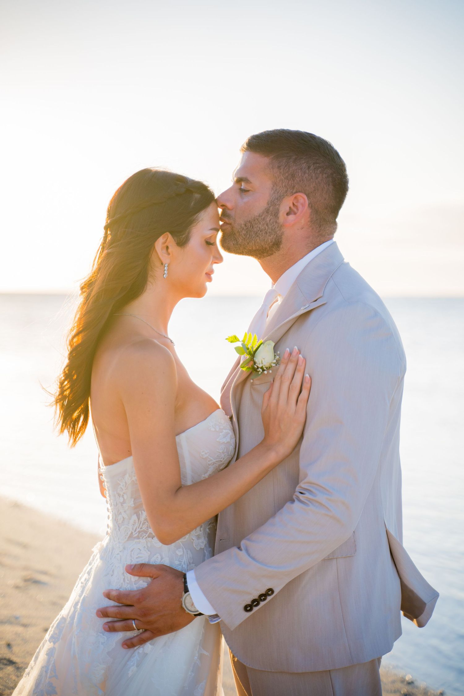 A couple in white attire walks hand in hand along a tropical beach with turquoise water and blue sky in the background.
