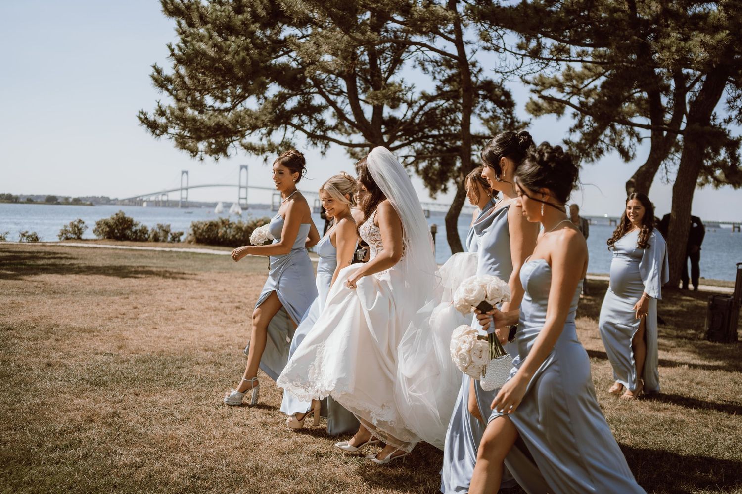 A wedding party in light blue and white dresses walk together on a grassy hill overlooking the ocean on a sunny day.