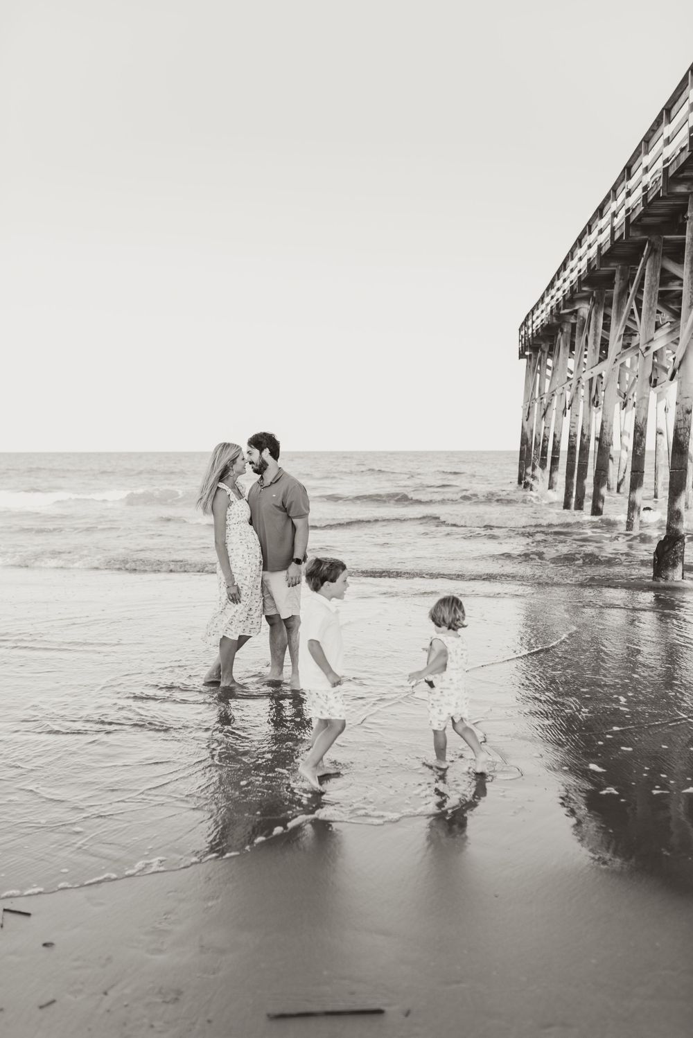 Family enjoying time together at beach near pier in black and white photograph.