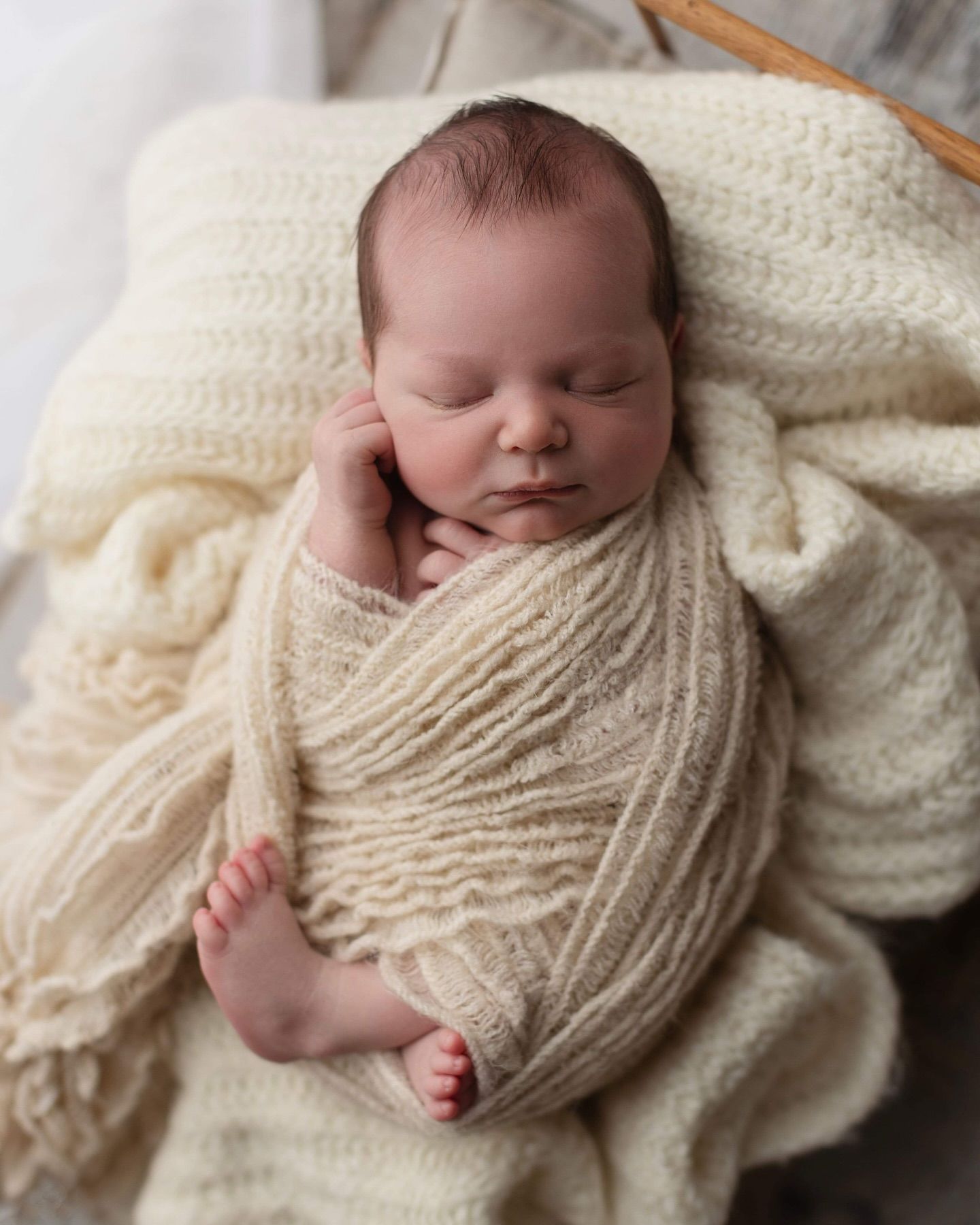 Sleeping newborn wrapped in soft cream-colored knit blanket with tiny feet peeking out against white textured background.