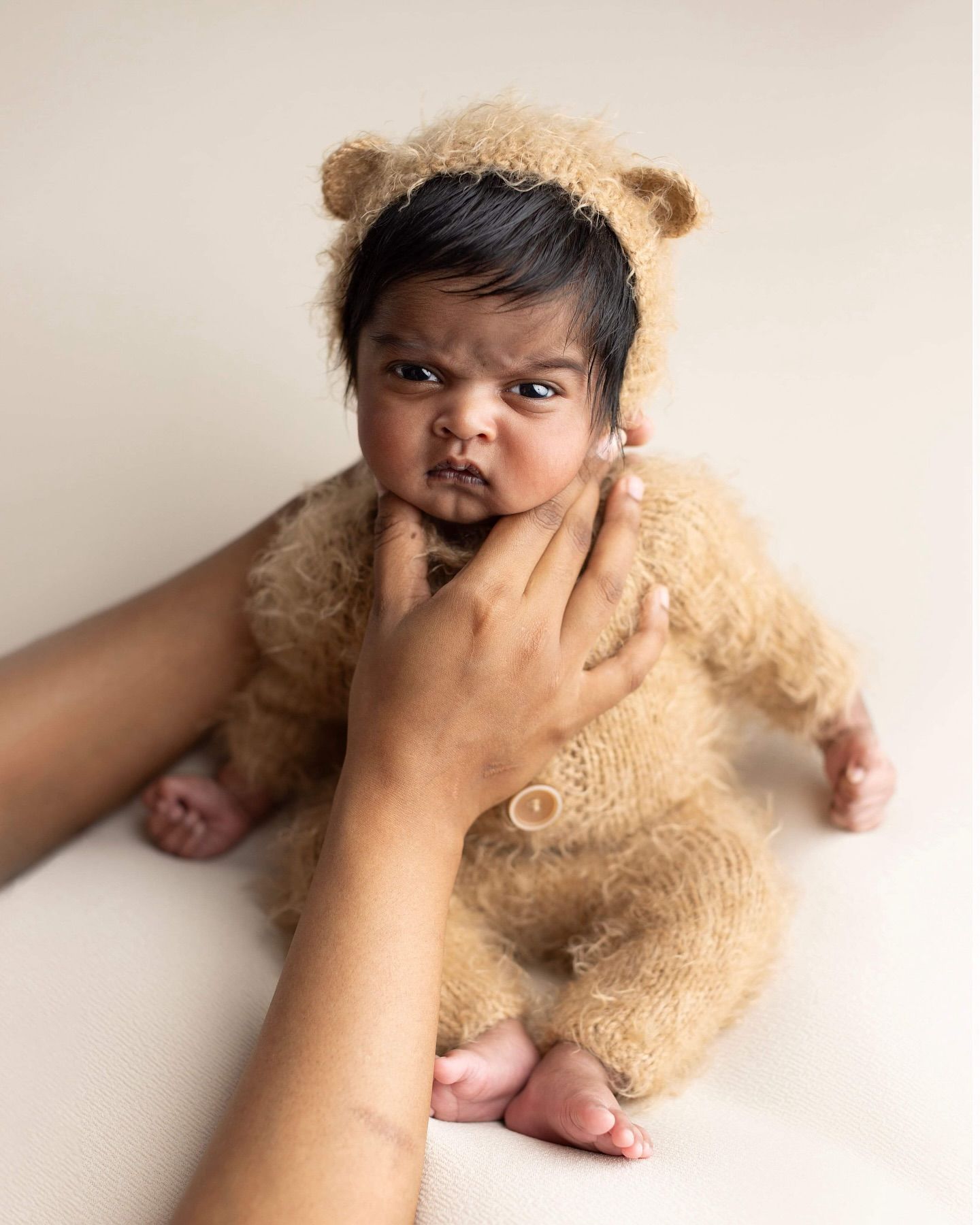 Baby wearing a plush teddy bear costume lies on a light background while a hand gently touches their cheek.