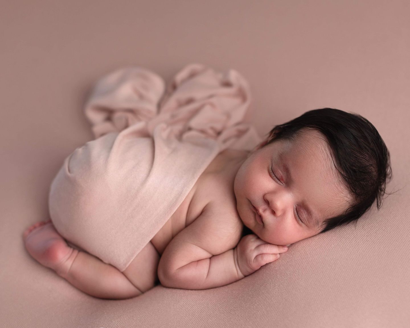 Sleeping newborn in pink wrap curled up peacefully on soft pink backdrop during professional photo session.