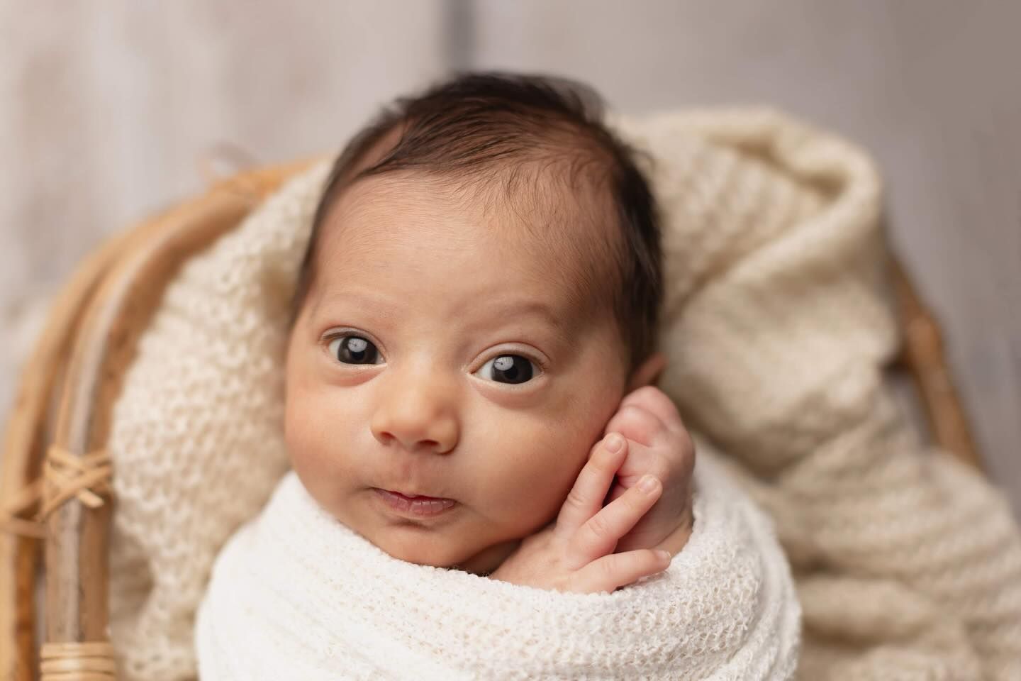 A newborn baby wrapped in a soft white blanket rests their head on their hand in a wooden basket.