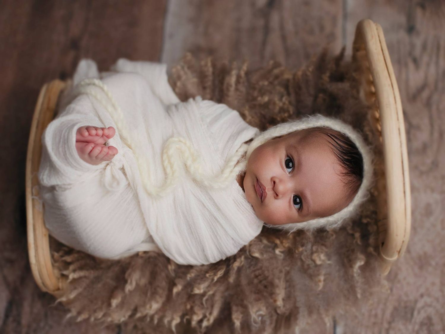 Sleeping newborn baby wearing white swaddle lying in wooden basket with brown fur lining.