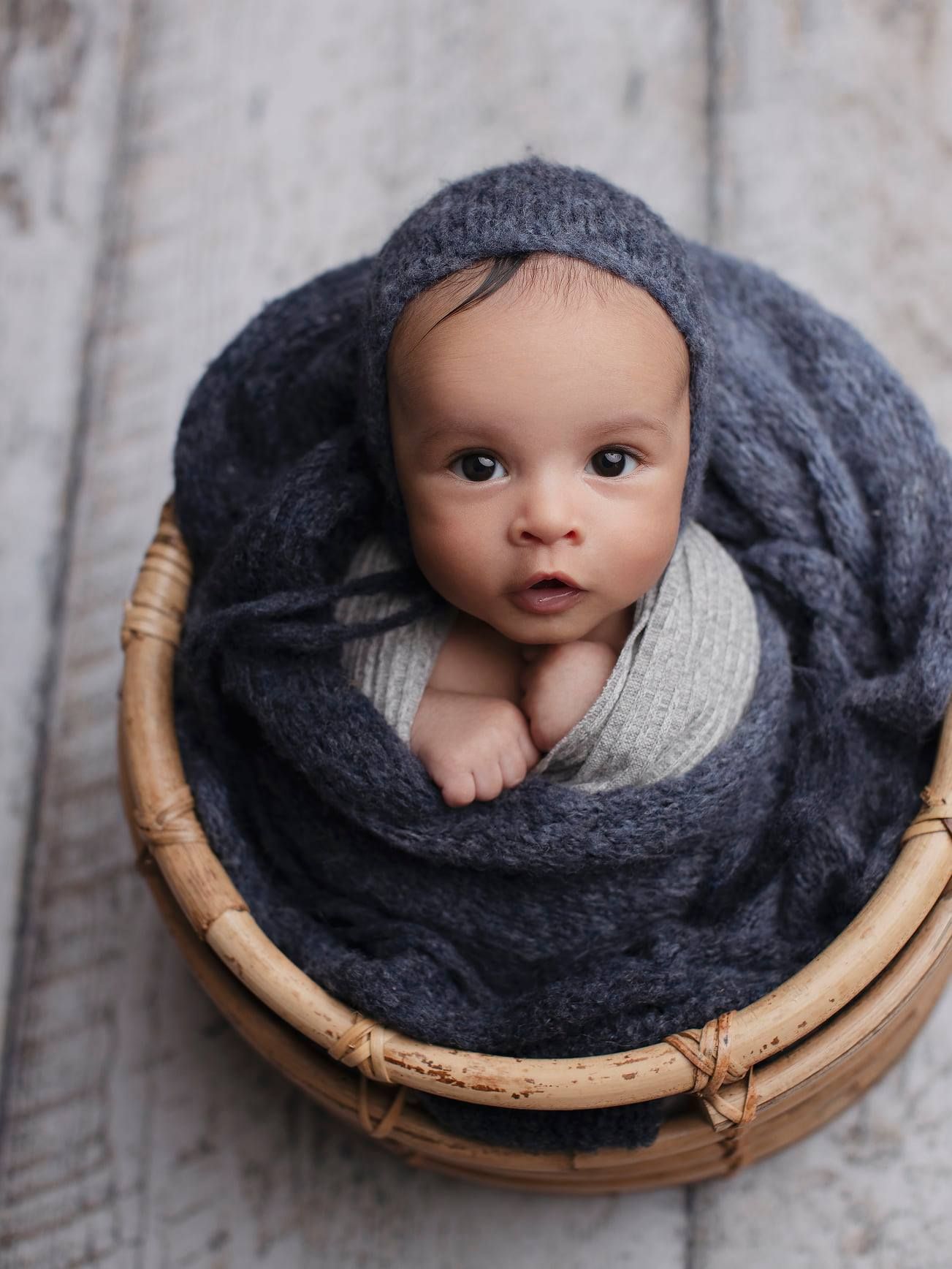 A small baby wrapped in a cozy gray knit blanket lies in a round wicker basket against a rustic wooden background.