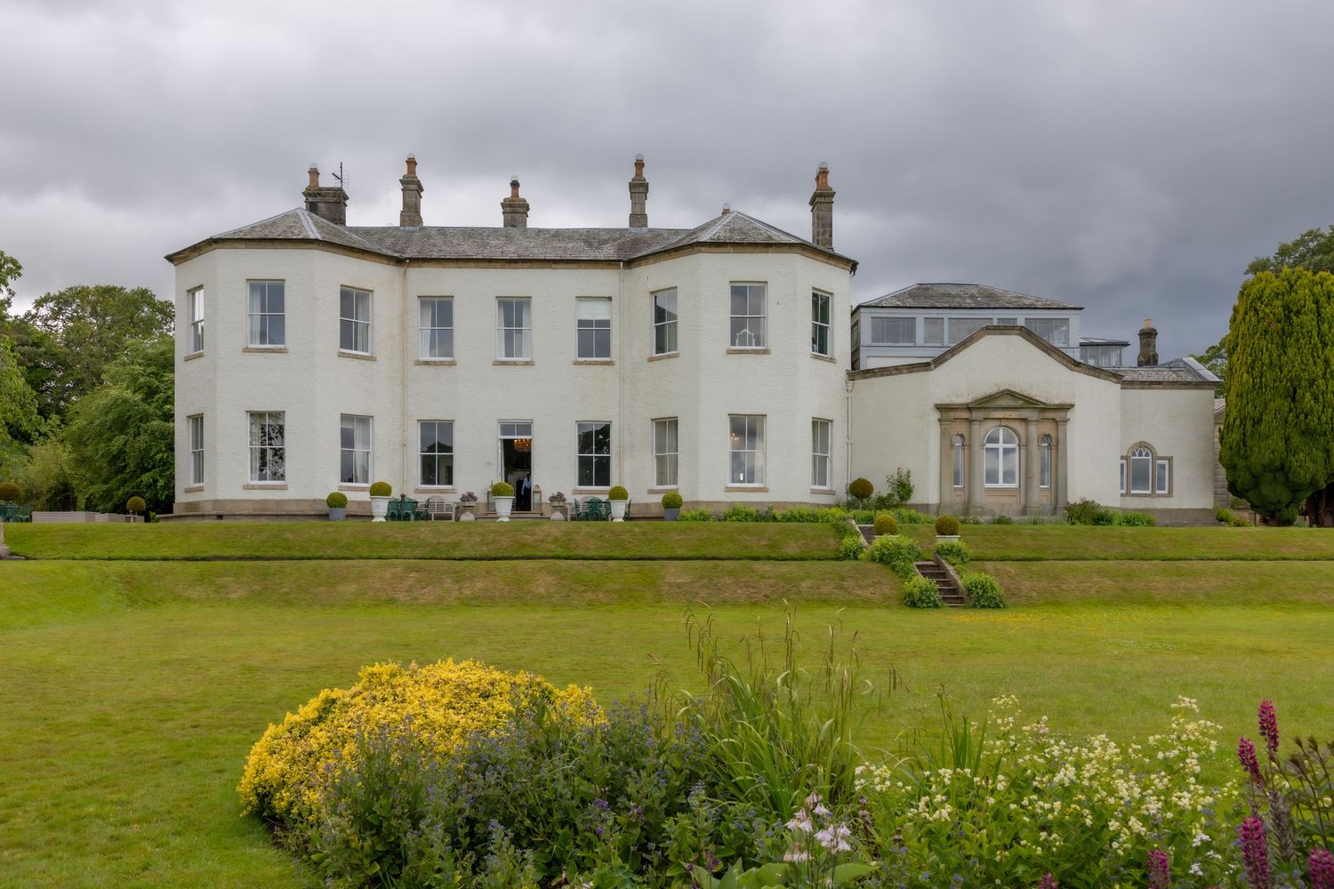 A white mansion with ornate architectural details sits atop a grassy hill surrounded by gardens and landscaping. Lartington Hall Wedding