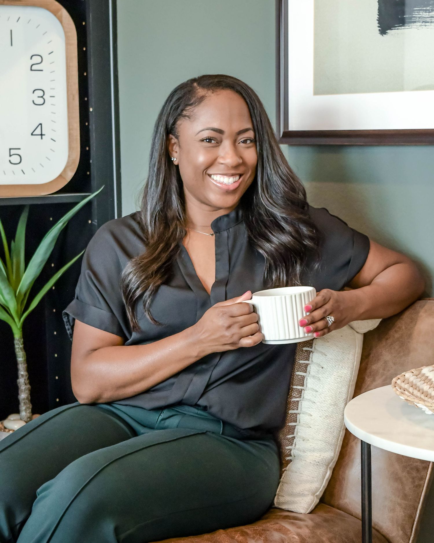 A woman relaxes on a leather couch with a coffee mug, next to a modern clock and plant in a cozy home setting.