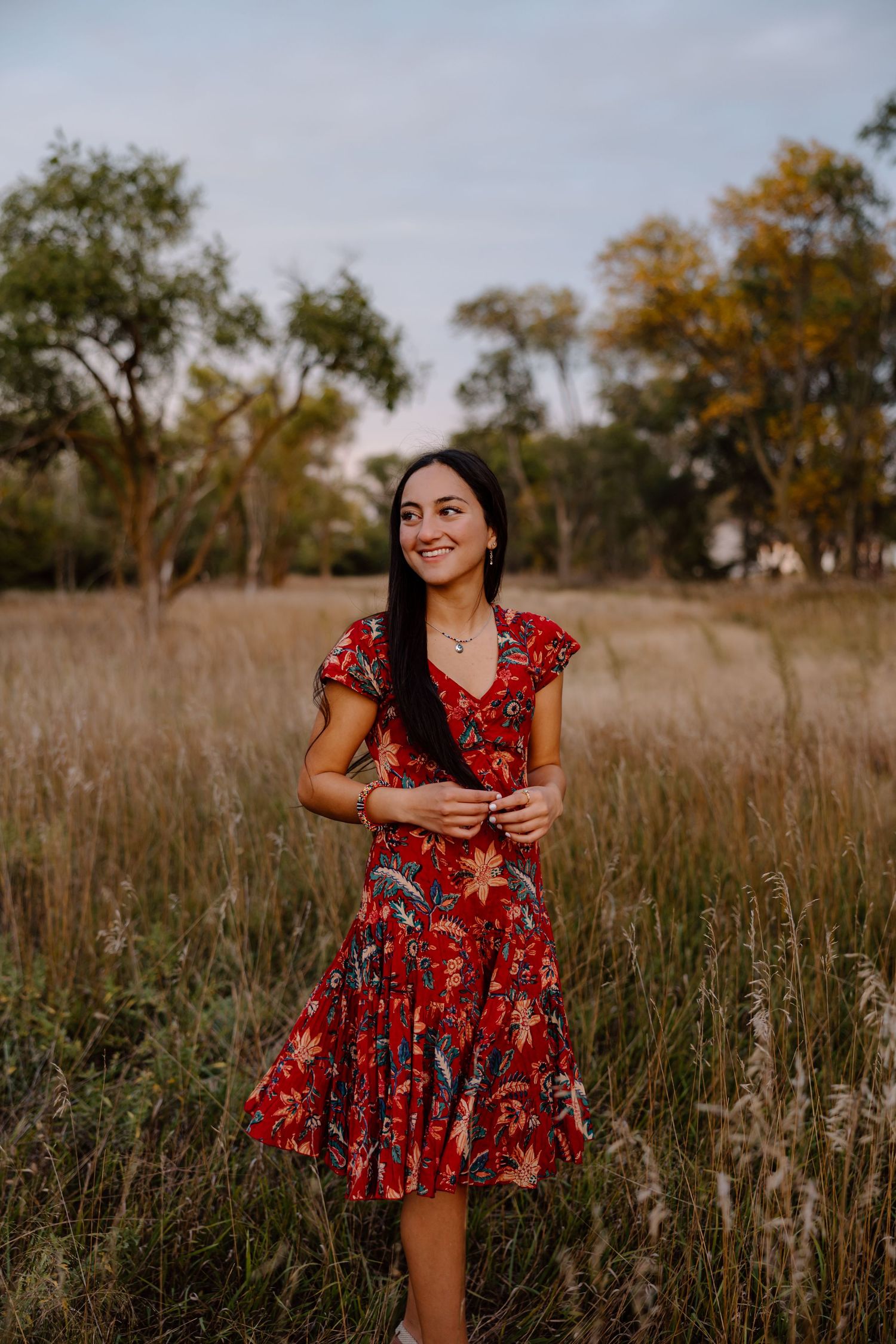 A person sits in a grassy meadow at sunset wearing a floral dress and glasses against a scenic waterfront backdrop.