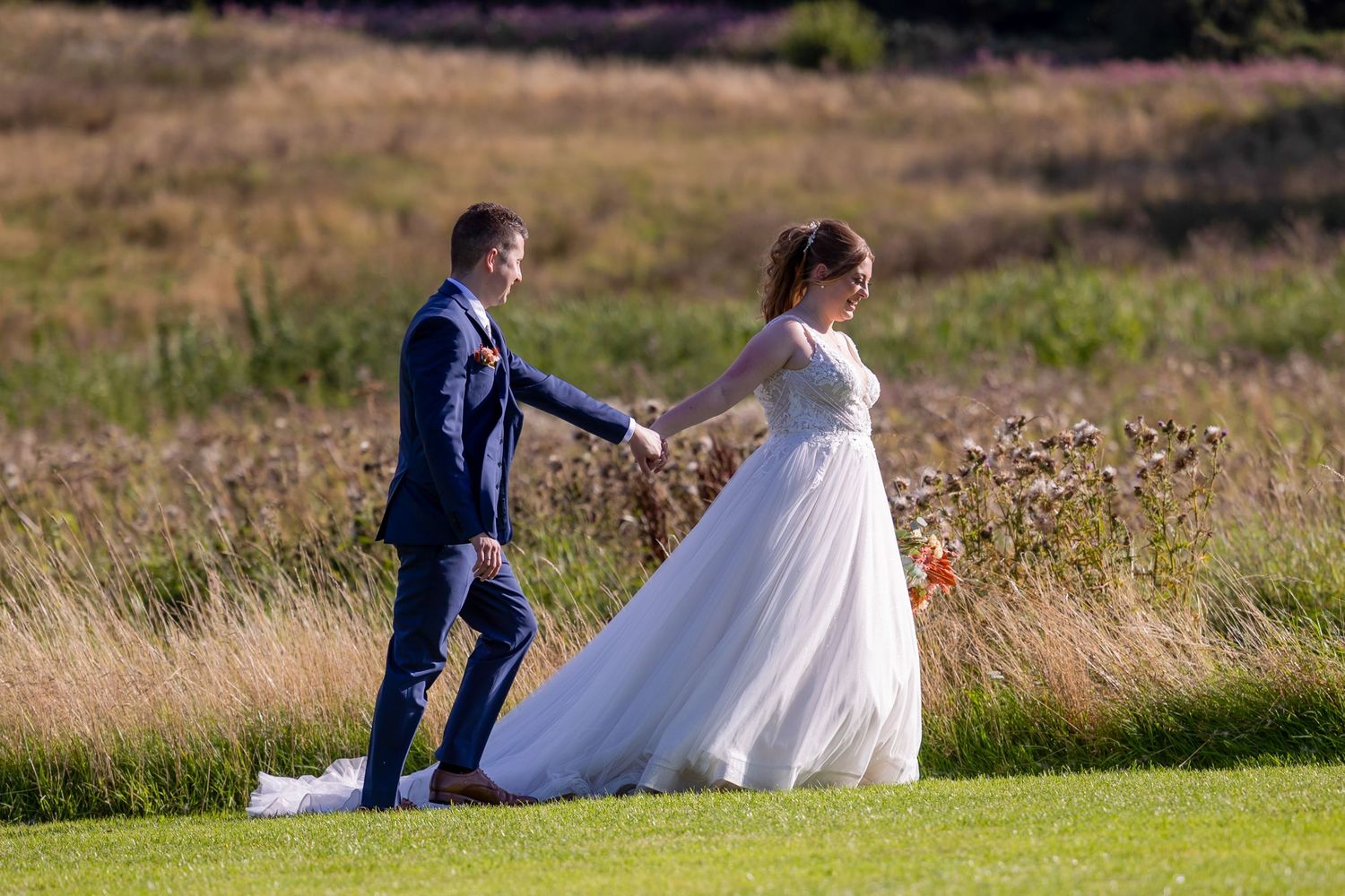A couple in wedding attire walks through a grassy field with birch trees in the background. South Causey Inn Farmhouse Wedding