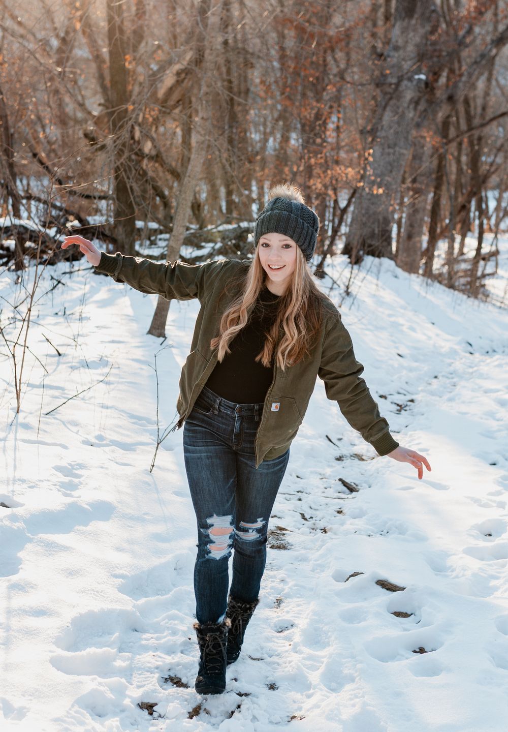 A person in winter clothing bends down to play in deep snow on a wooded trail during a winter day.