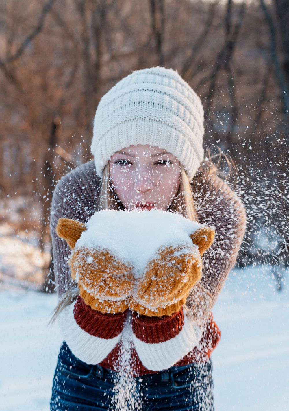 Someone wearing cozy winter mittens and a white knit hat blows fresh snow into the air on a cold winter day.
