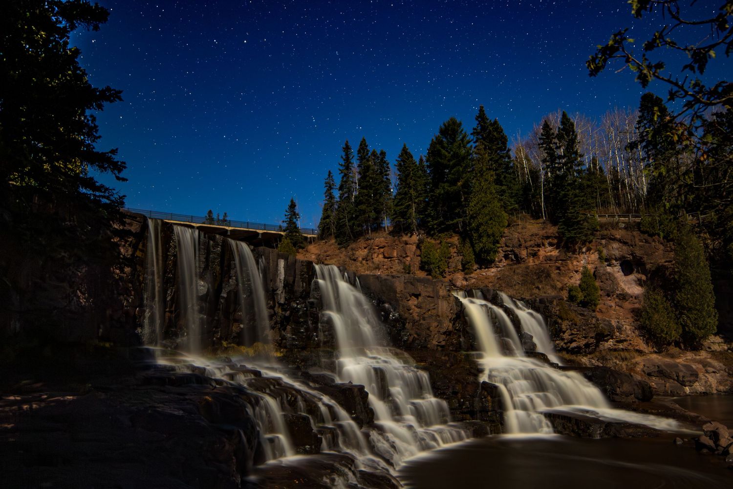 Night sky landscape photography of Gooseberry Falls State Park beneath star-filled sky on a full moon night, dramatic celestial wall art.