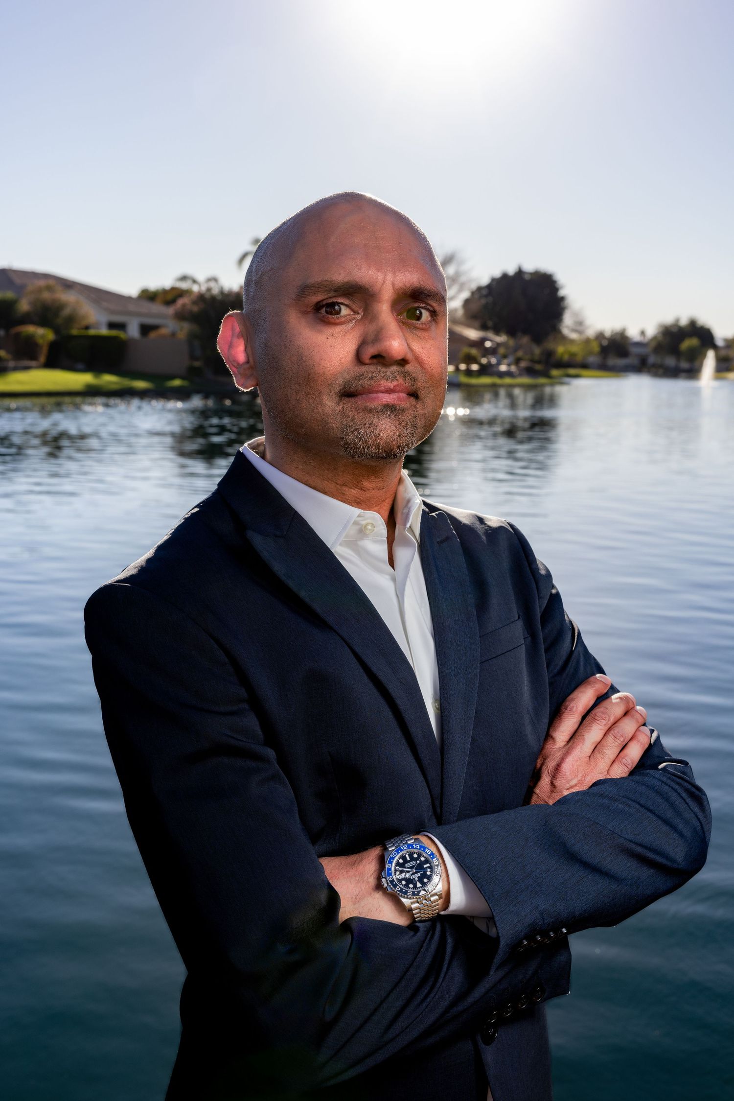 Professional man in navy blazer stands confidently with arms crossed beside a serene lake at golden hour.