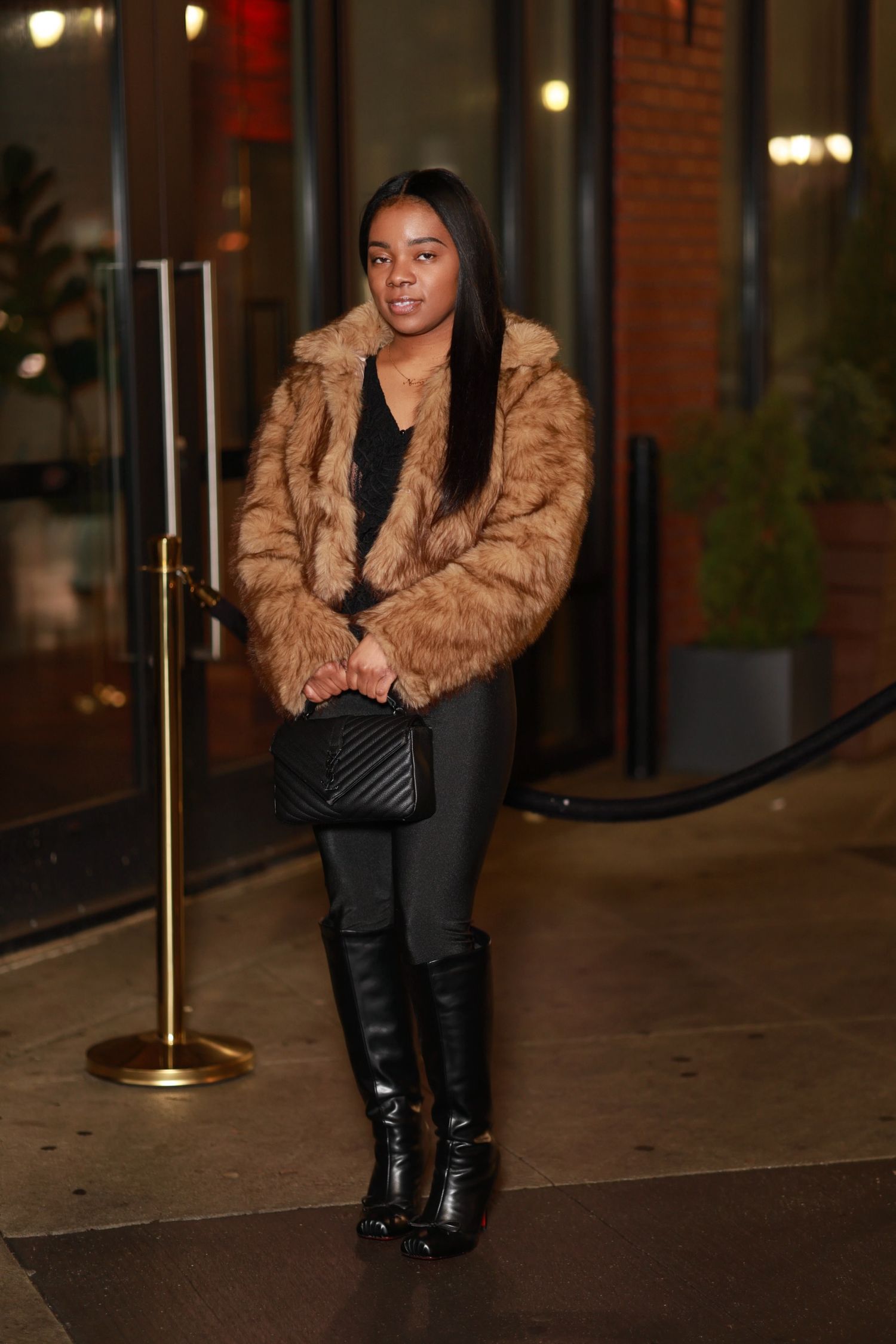 Woman in brown fur coat and black boots poses outside building entrance with gold stanchions at night.