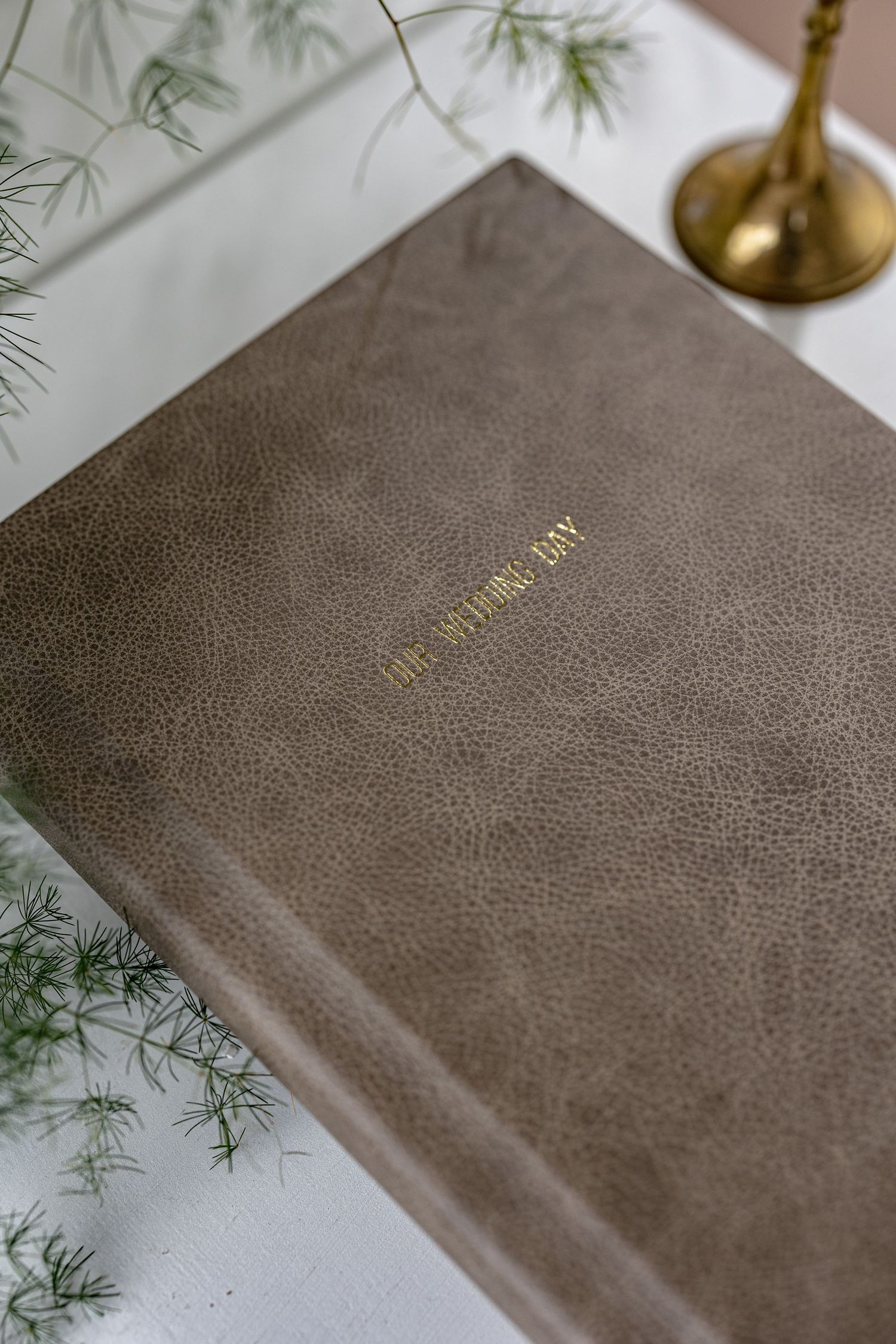 A brown leather-bound book or journal with gold text on the cover sits on a white surface near delicate green foliage.