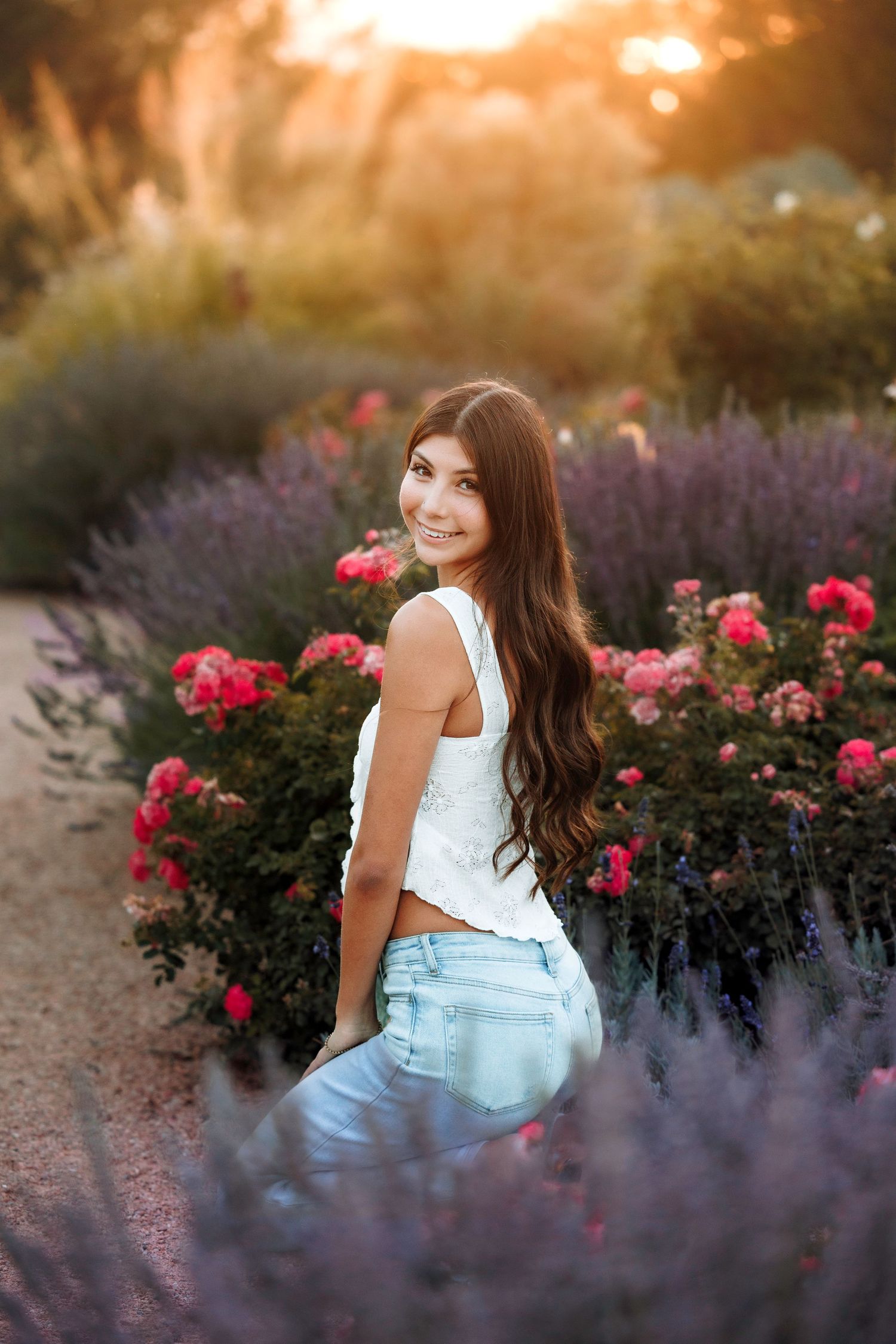 Young woman with long dark hair smiling in a garden with pink roses and lavender at golden hour.