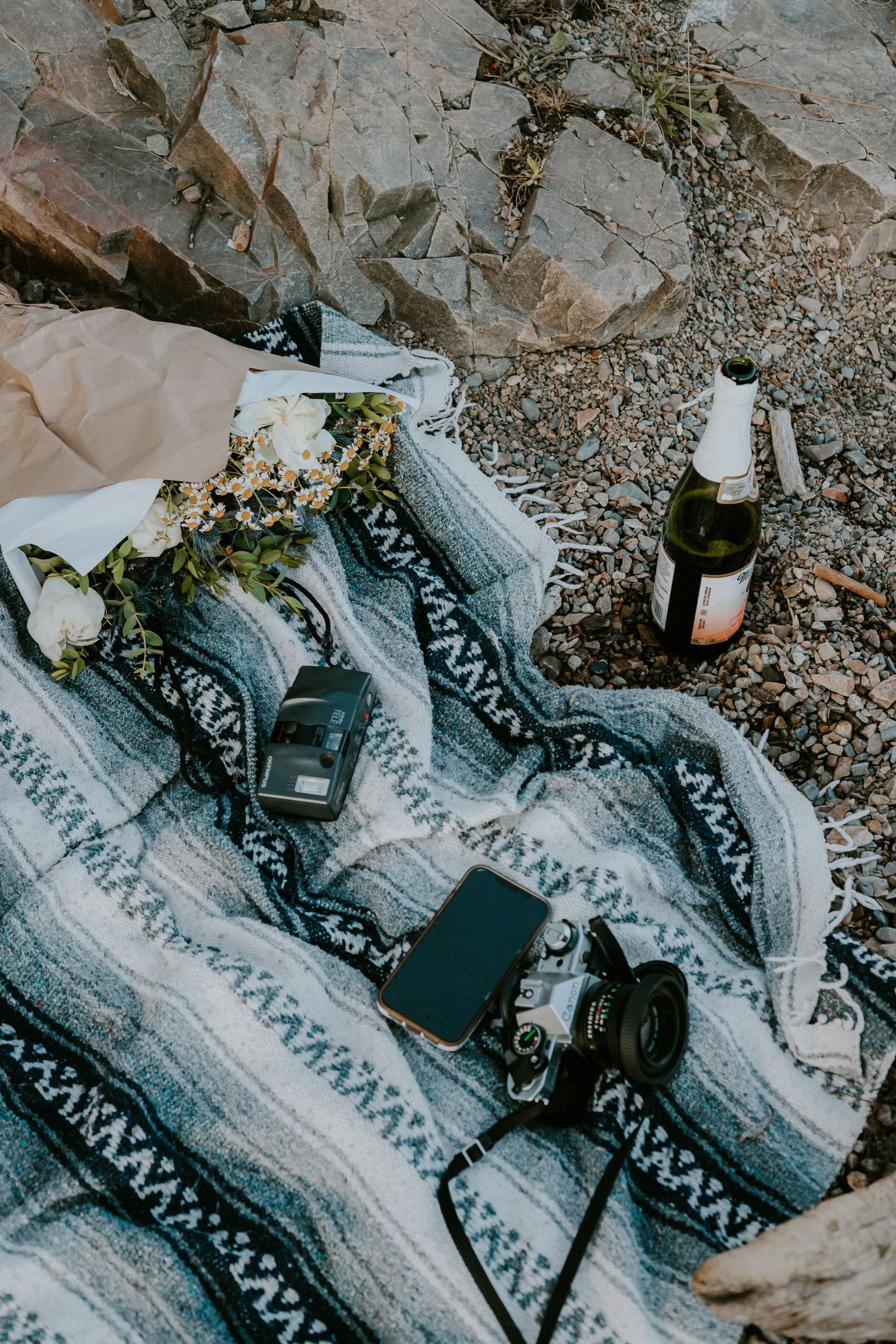 Vintage cameras and wine bottle arranged on patterned blanket against rocky backdrop.