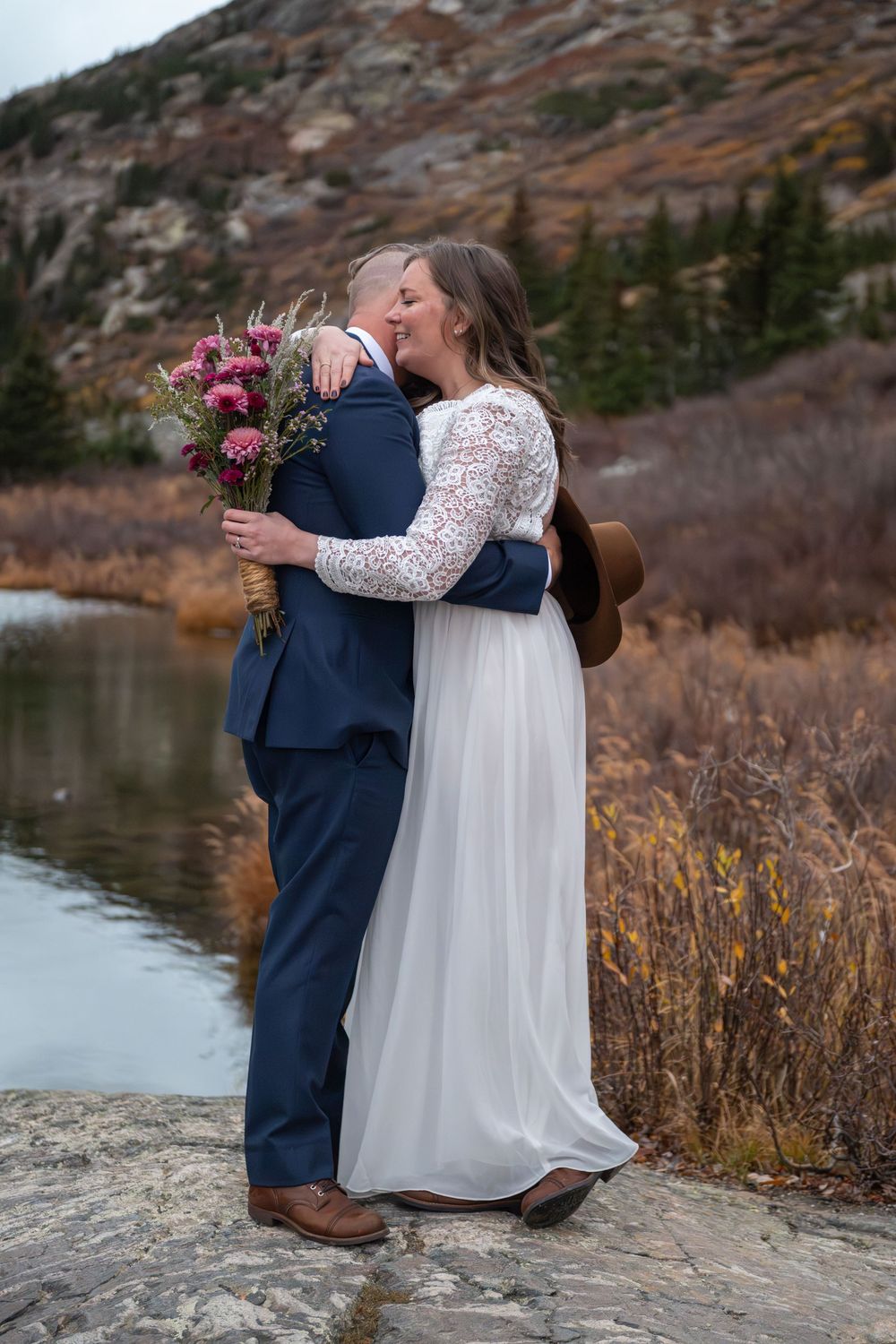 An eloping couple share a an intimate embrace by 
a high mountain lake with rugged peaks surrounding them.