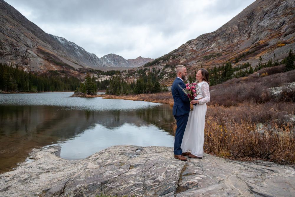 An eloping couple share a quiet moment together on the edge of an alpine like with mountains all around the. She is in a white dress.