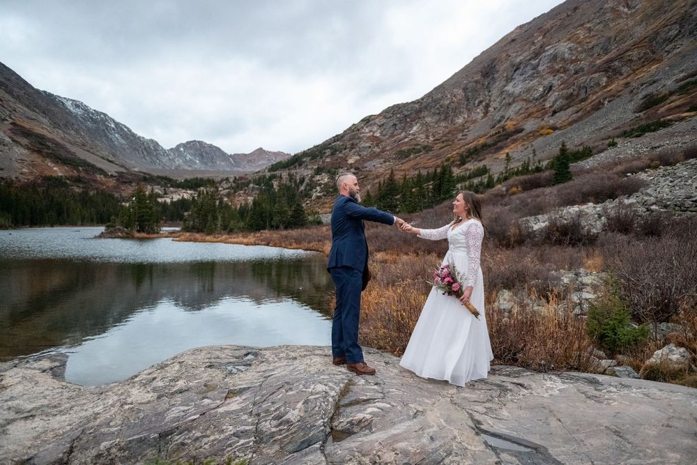 An eloping couple toast with coffee at sunrise by a high mountain lake with rugged peaks surrounding them.
