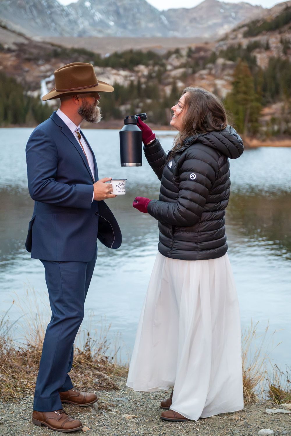 A bride and groom share coffee at sunrise by a high mountain lake with rugged peaks surrounding them.